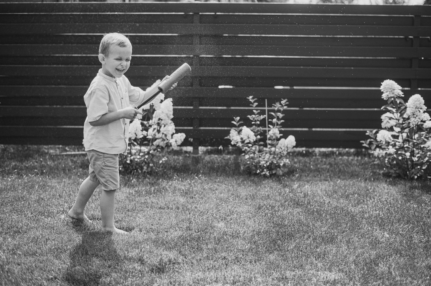 Black and white photo of young boy swinging a bat in a backyard garden.