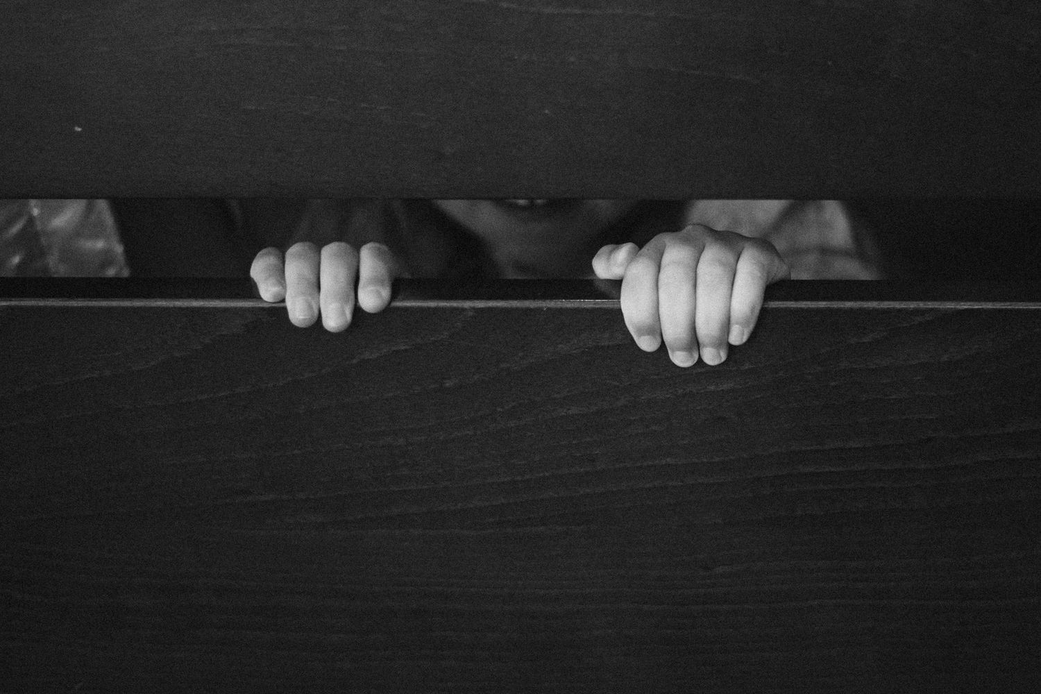 Child peeking over dark wooden surface in moody black and white photo.