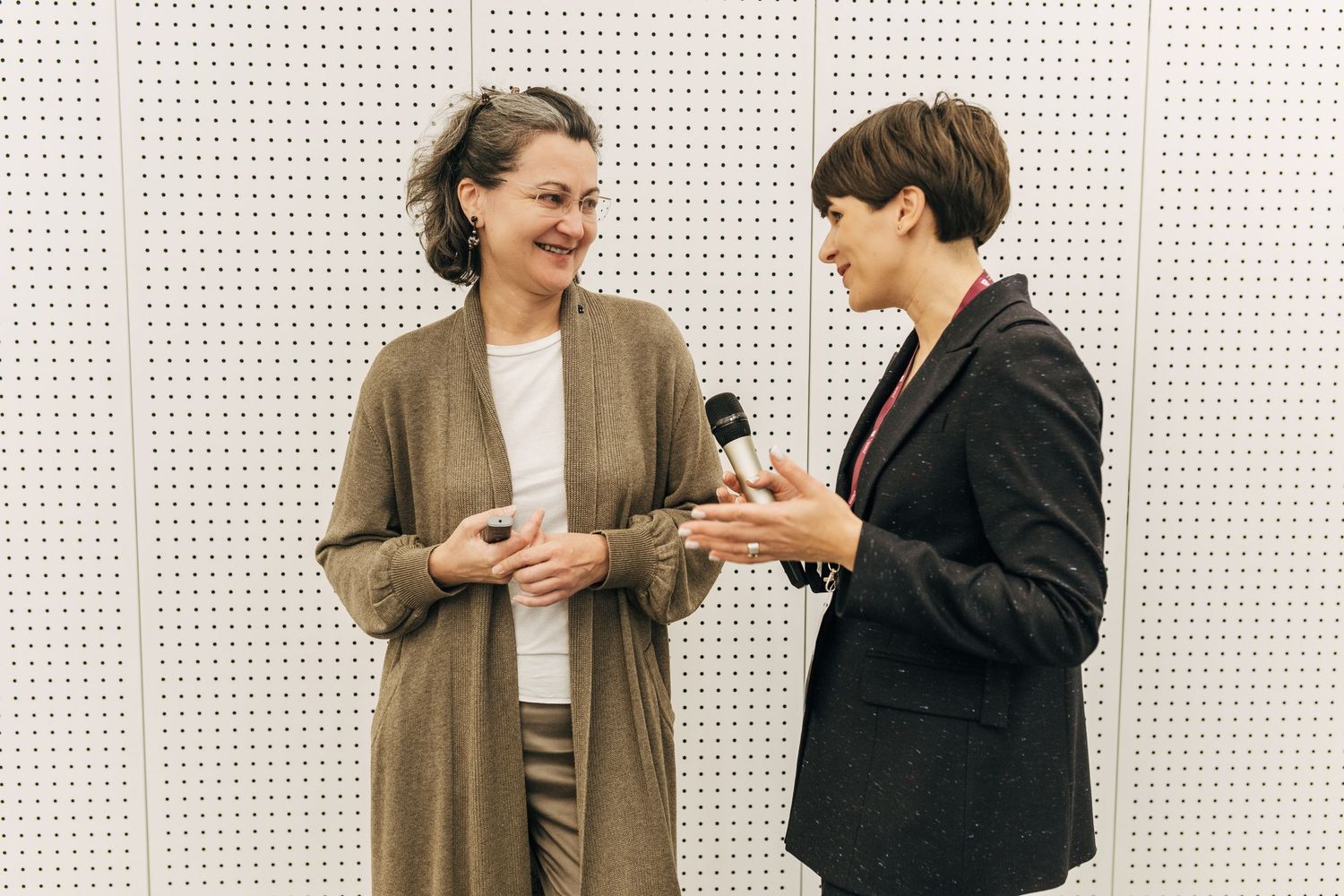 Two people share a laugh while holding beverages during a professional event in front of a dotted wall backdrop.