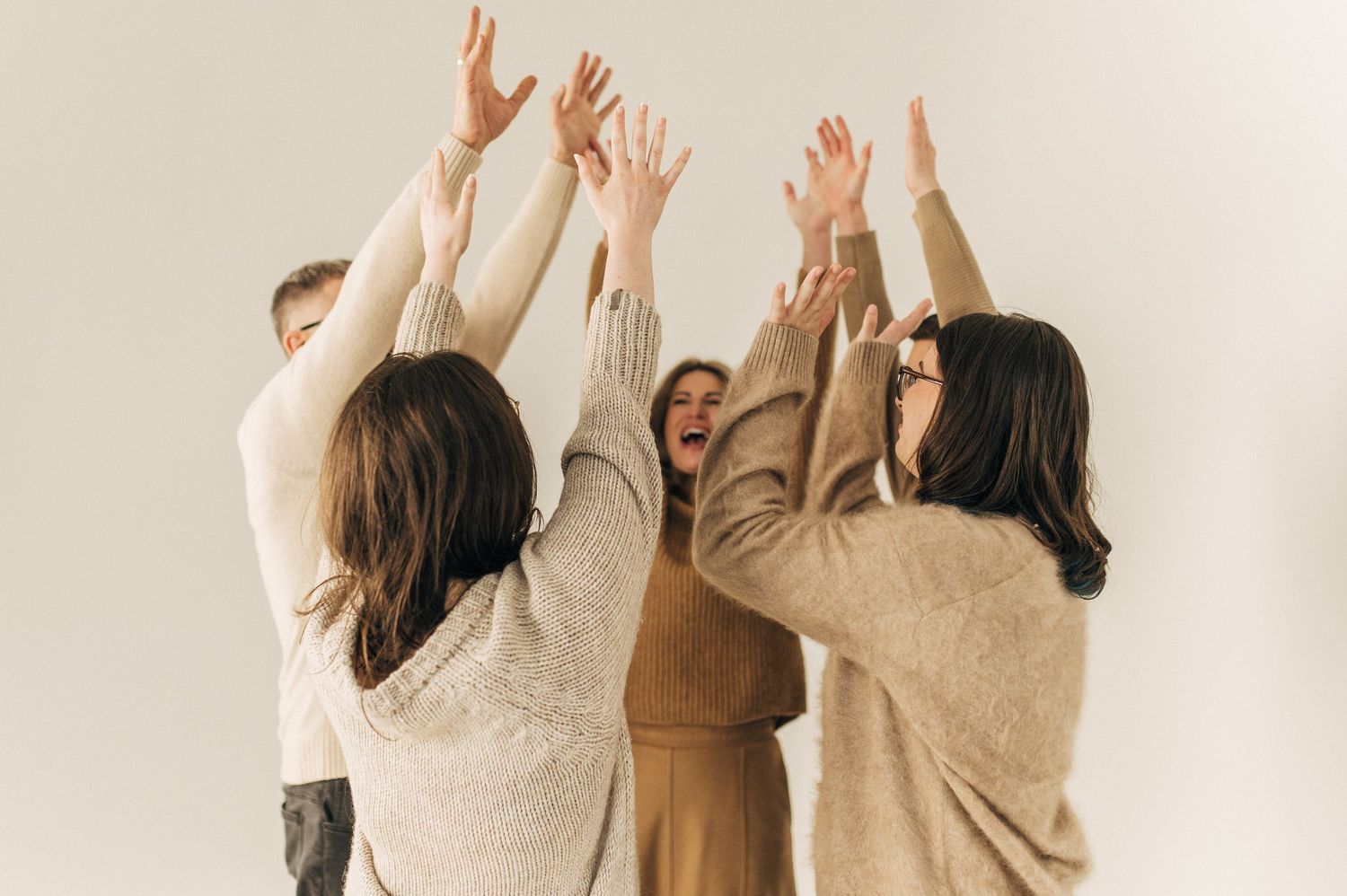 Group of people with raised hands wearing neutral colored clothing against a white background.