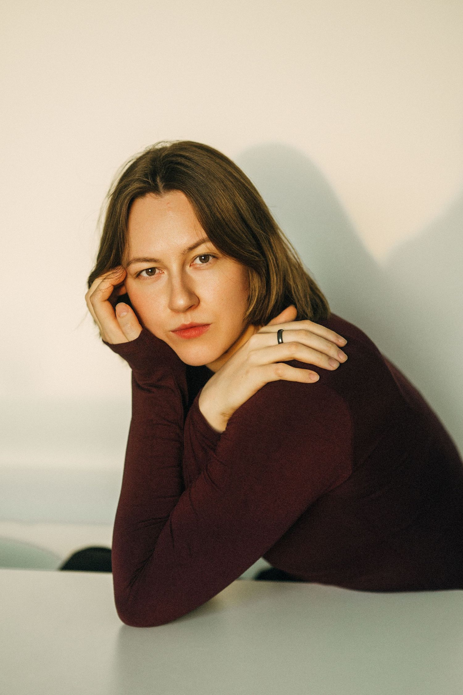 Young woman in burgundy turtleneck resting chin on hand at white table, gazing thoughtfully at camera.