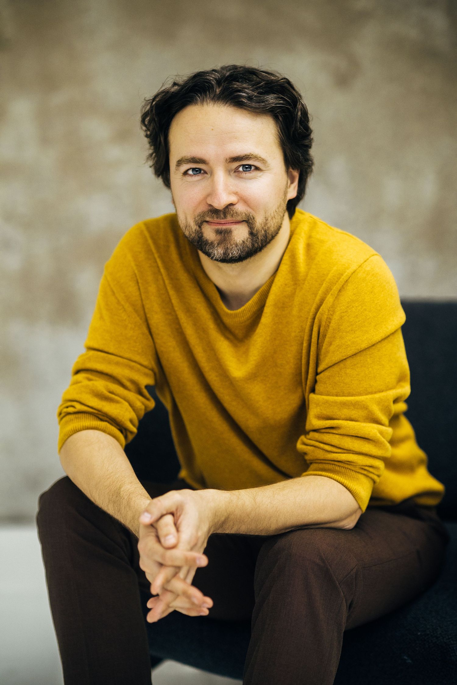 A smiling bearded man in a yellow sweater sits relaxed with hands clasped against a neutral gray background.