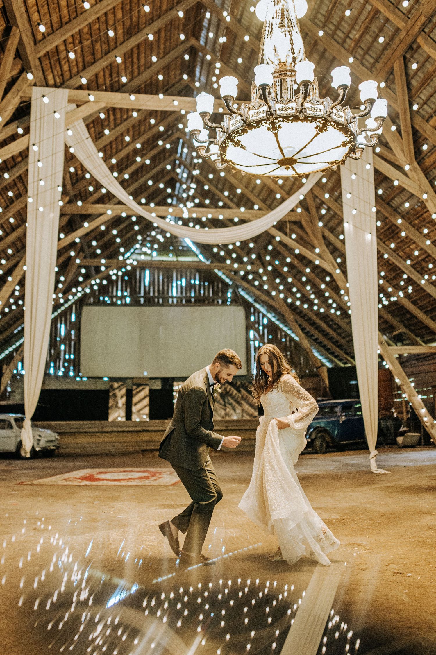 A couple dances beneath twinkling lights and a chandelier in a rustic barn wedding venue.