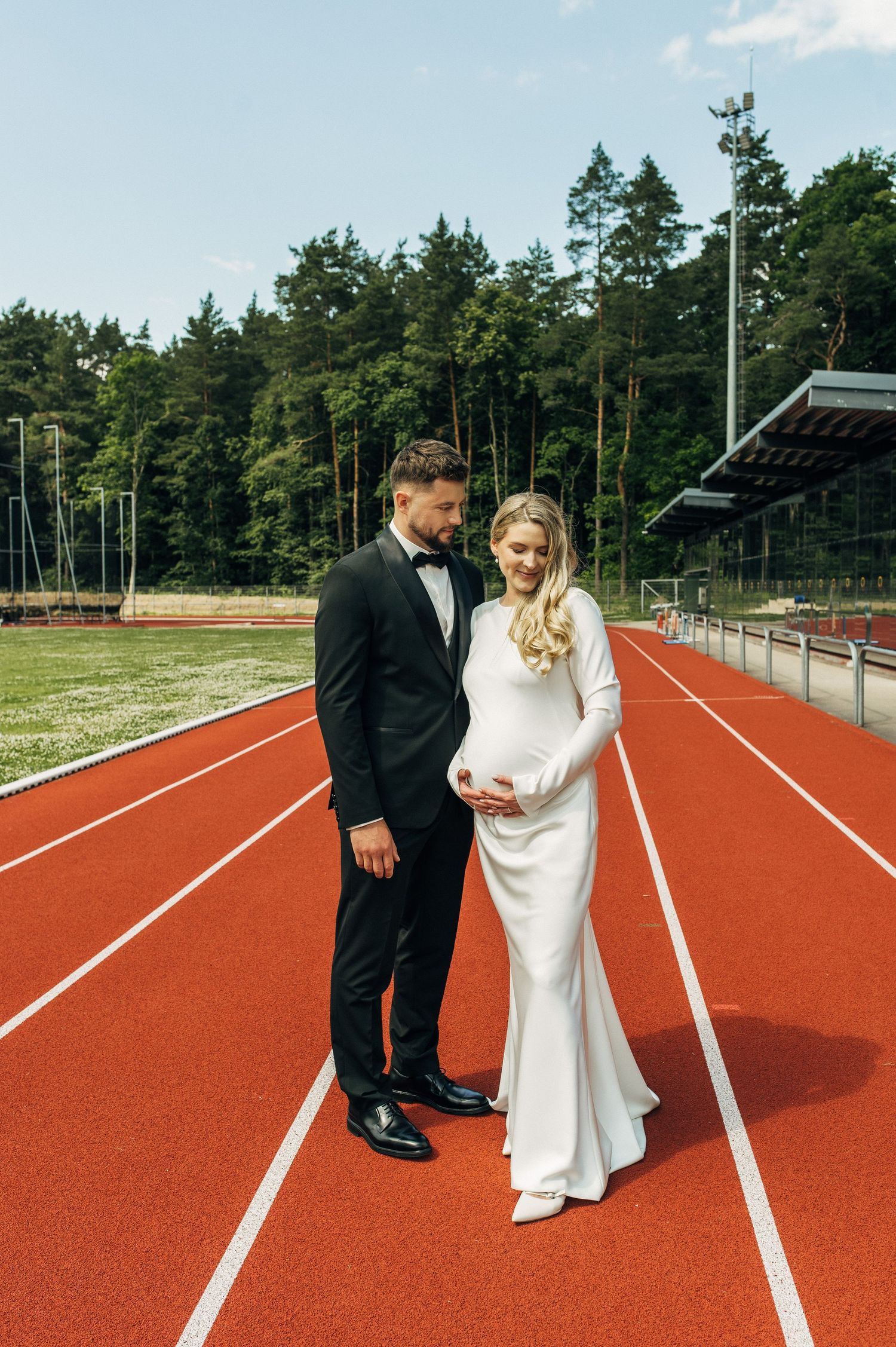Pregnant bride in white gown and groom in black tuxedo standing on red running track with forest backdrop.