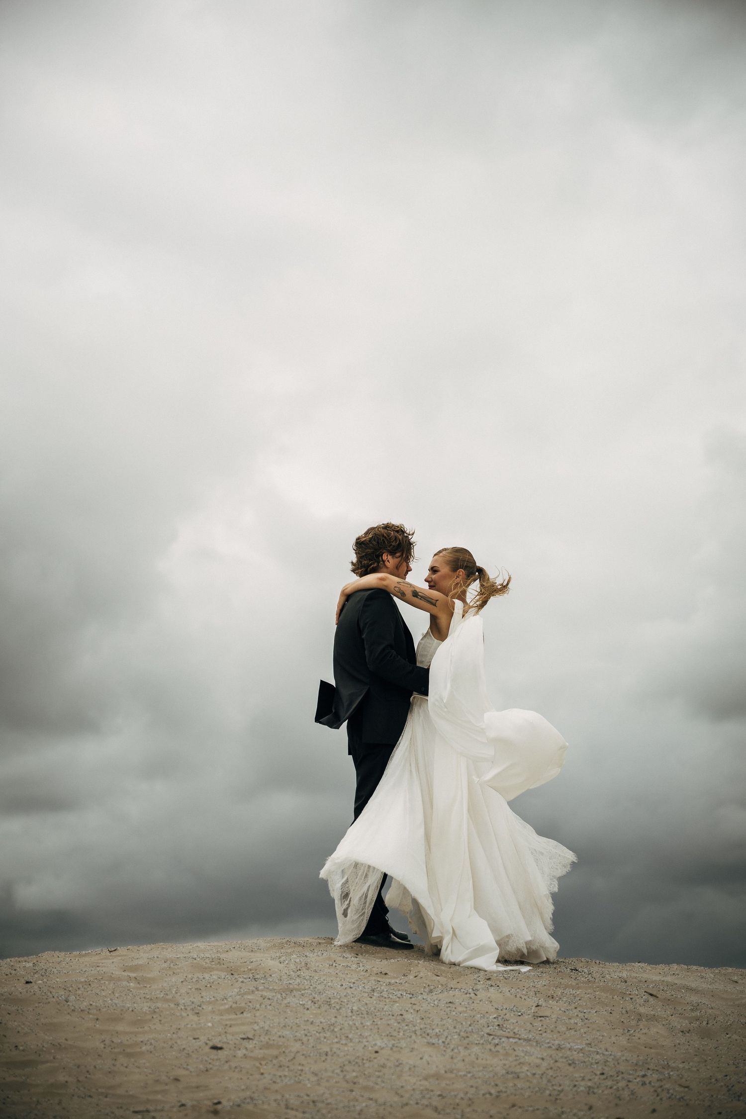 Bride and groom embracing on sandy dunes under dramatic stormy sky, wedding dress flowing in wind.