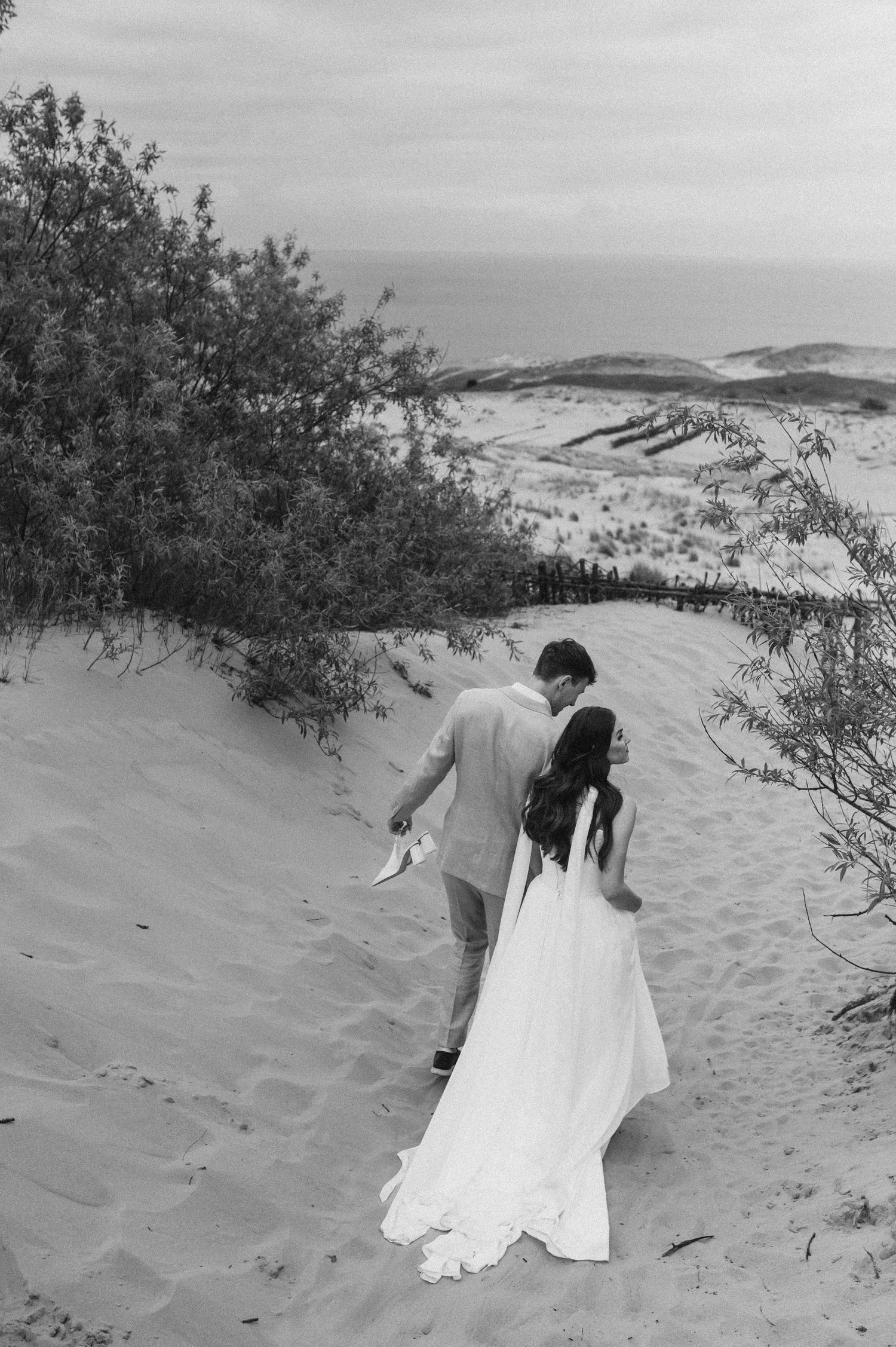 A couple in formal attire walks together on sand dunes with beach grass and rolling hills in the background.