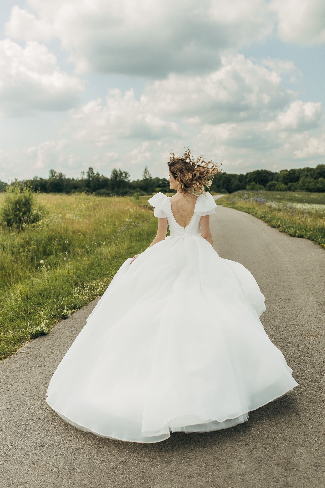 White wedding dress flowing on a country path surrounded by green meadows and summer skies.