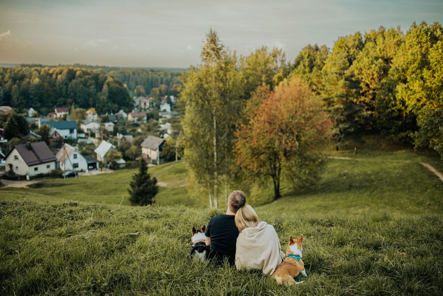 A couple and their dog sit together on a grassy hillside overlooking a small village.