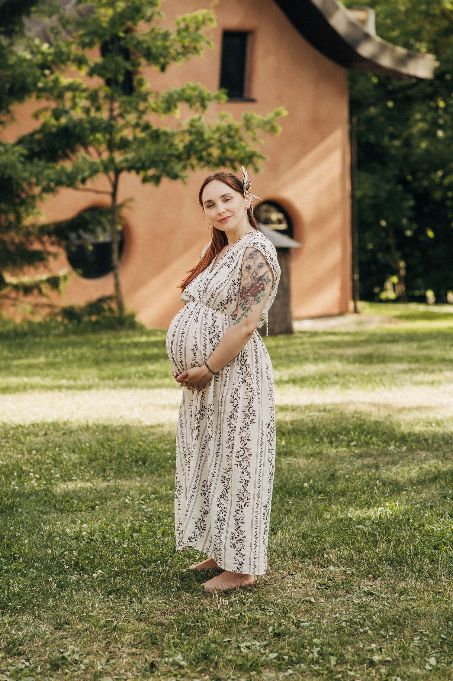 Pregnant woman in patterned dress stands in garden with rustic building in background.