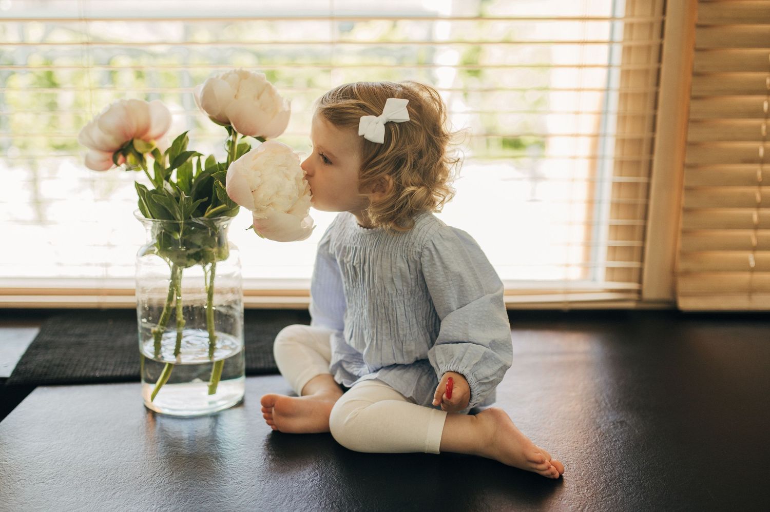 Child sitting at dark table with vase of flowers in sunlit room.