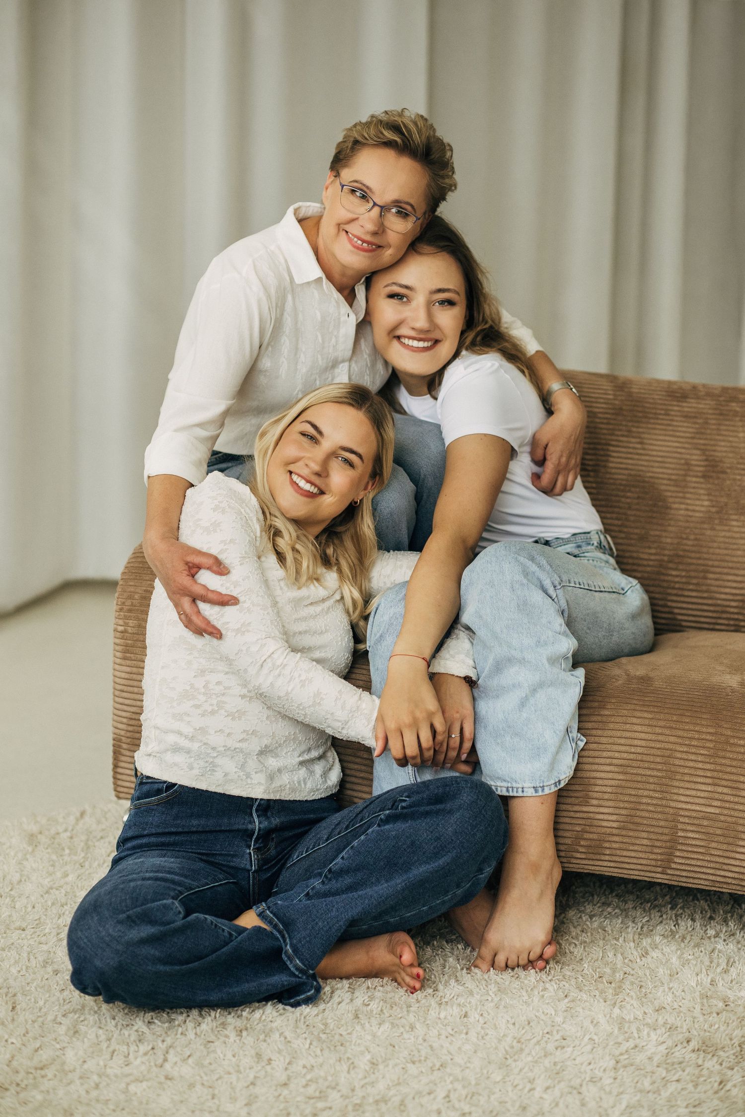 Three smiling women pose together on a couch at home, sharing a warm embrace in a cozy living room.