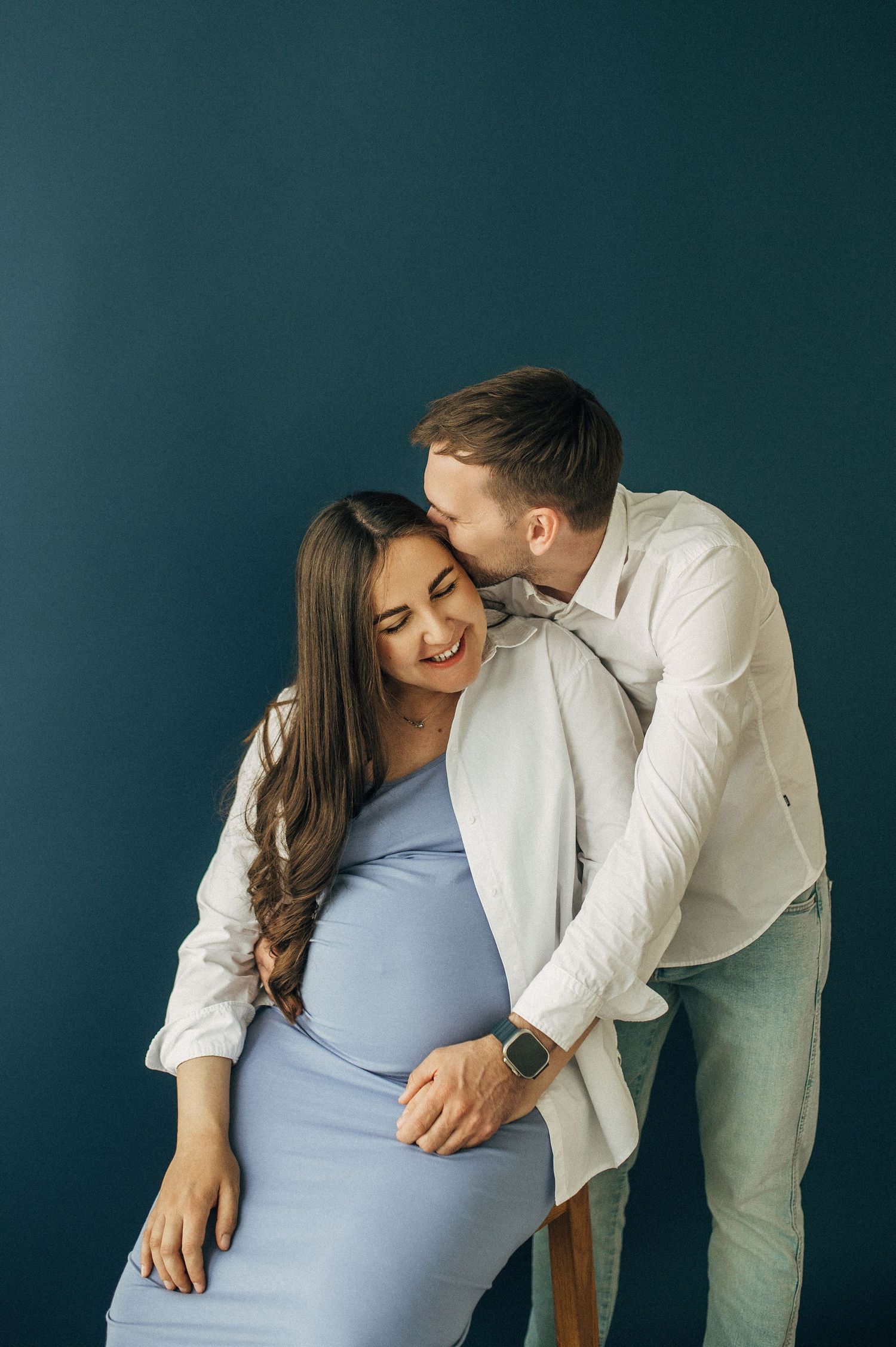 Intimate maternity portrait with couple in white and blue against dark backdrop.