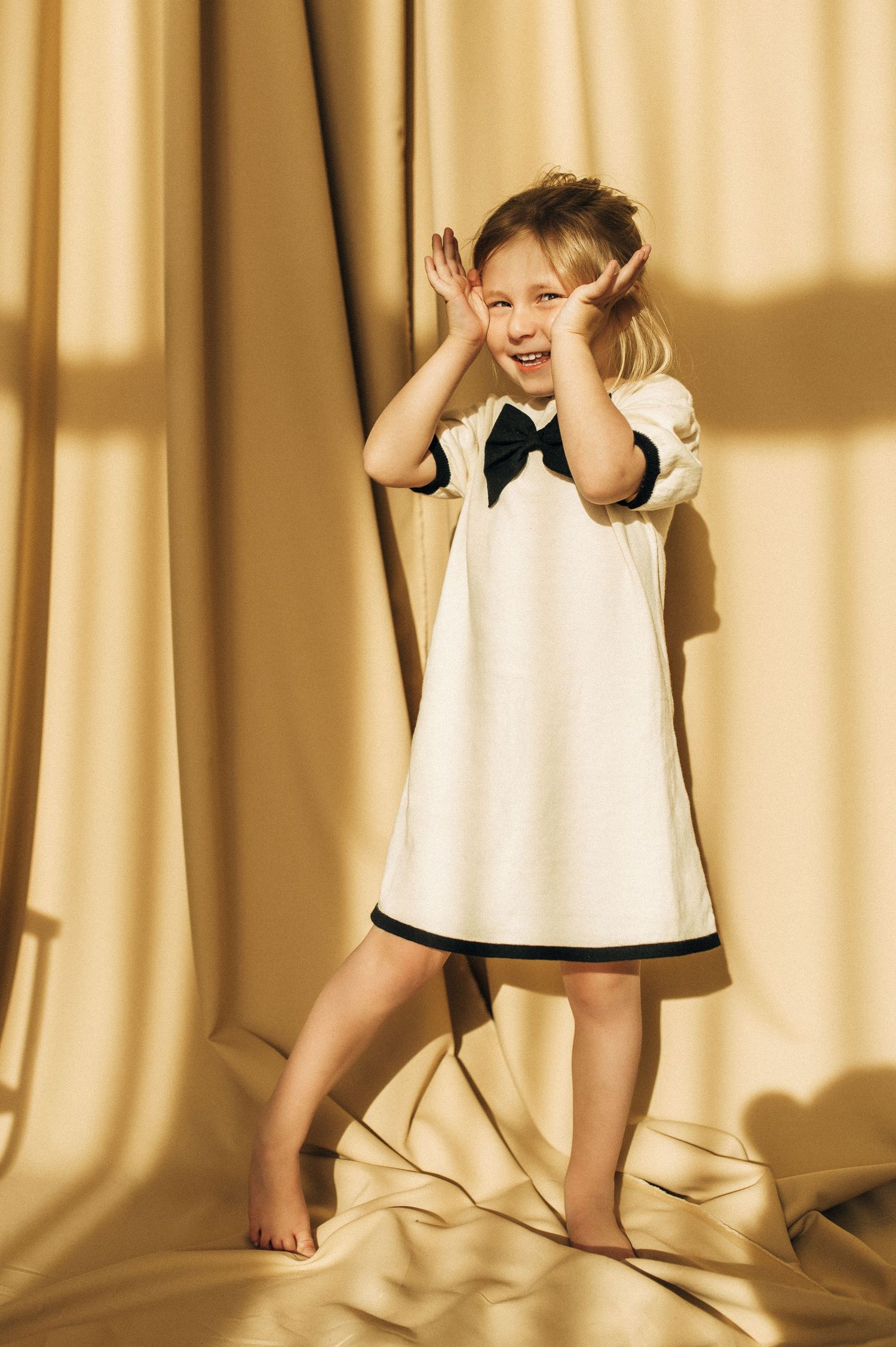 Playful child wearing white dress poses against golden sunlit backdrop.