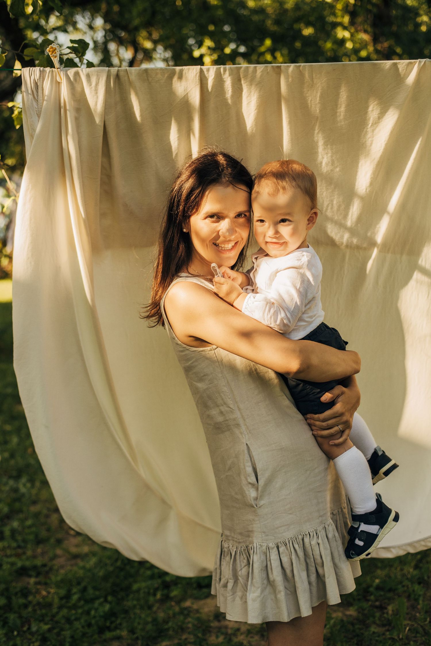 Smiling mother in linen dress holding baby boy outdoors in golden sunlight.