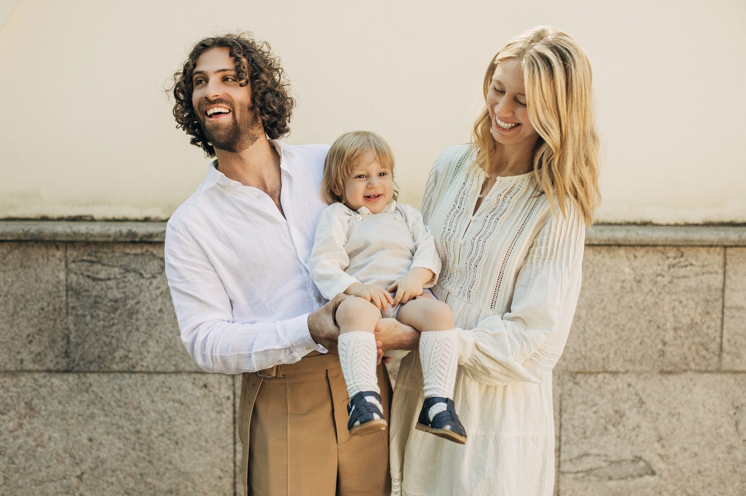 Happy young family with curly-haired father, blonde mother, and toddler posing outdoors.