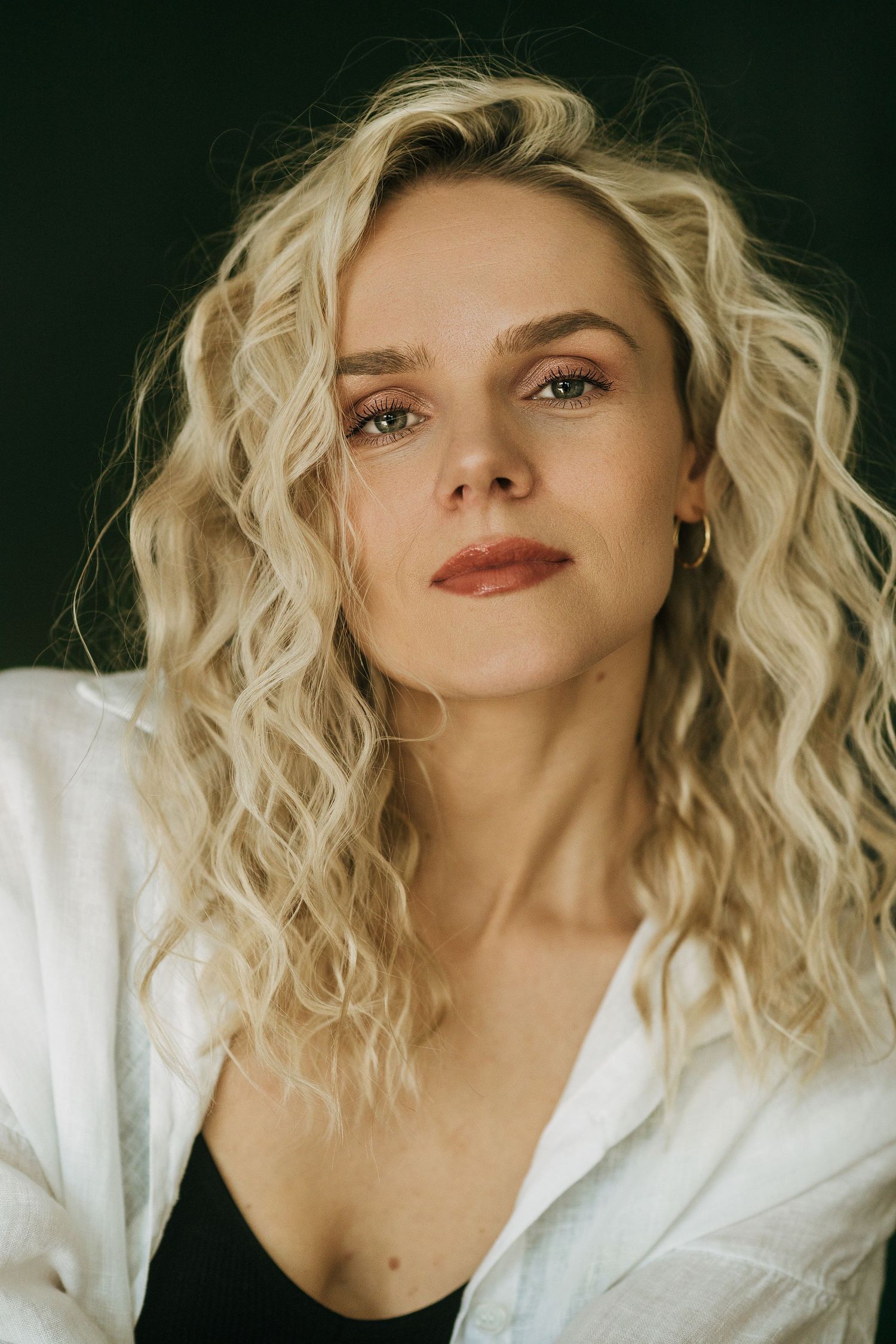 Portrait of a young woman with curly blonde hair, green eyes, and a subtle smile wearing a white shirt.