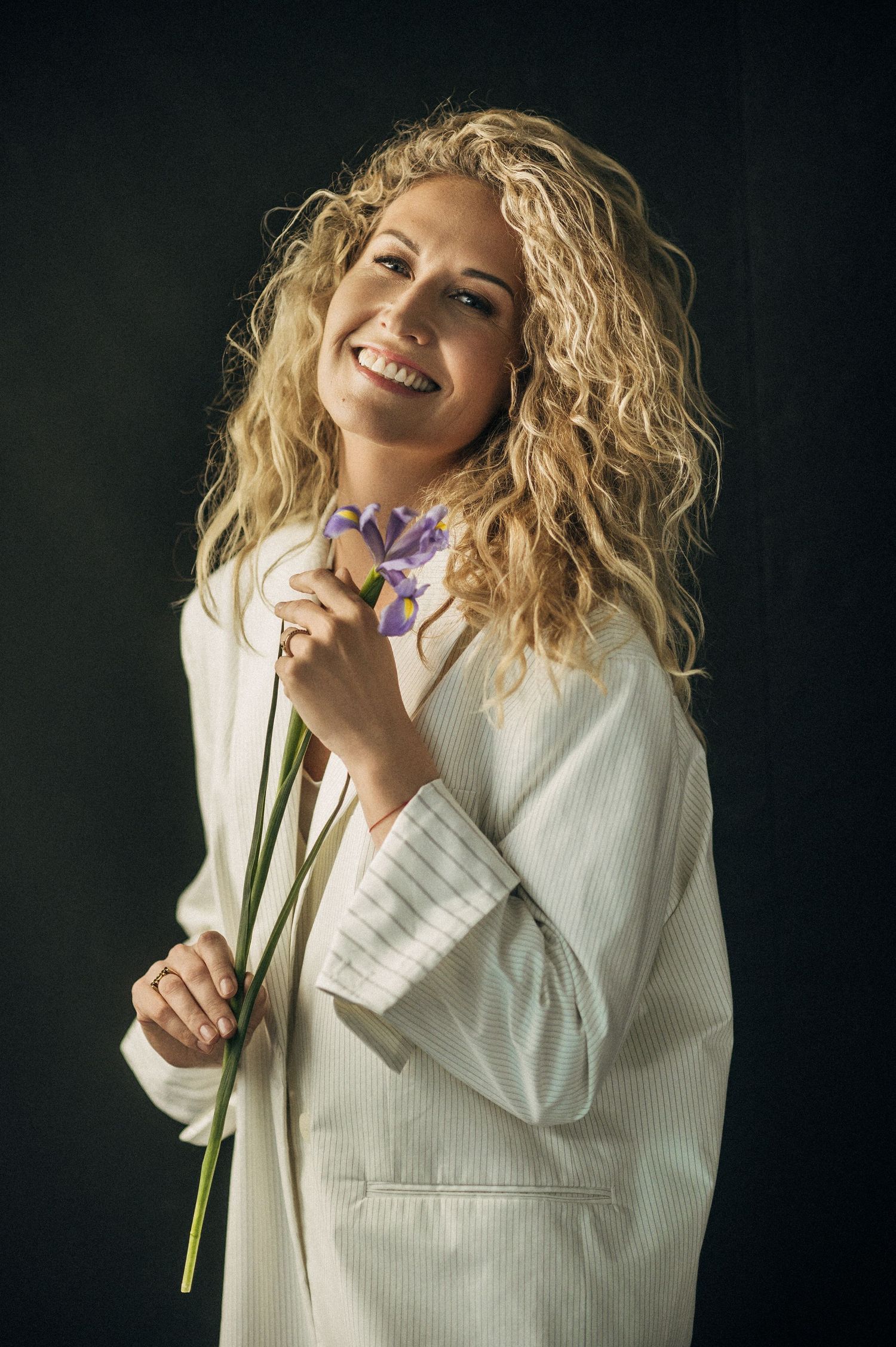 Smiling blonde woman with curly hair holding a purple iris flower, wearing a white blazer against a dark background.