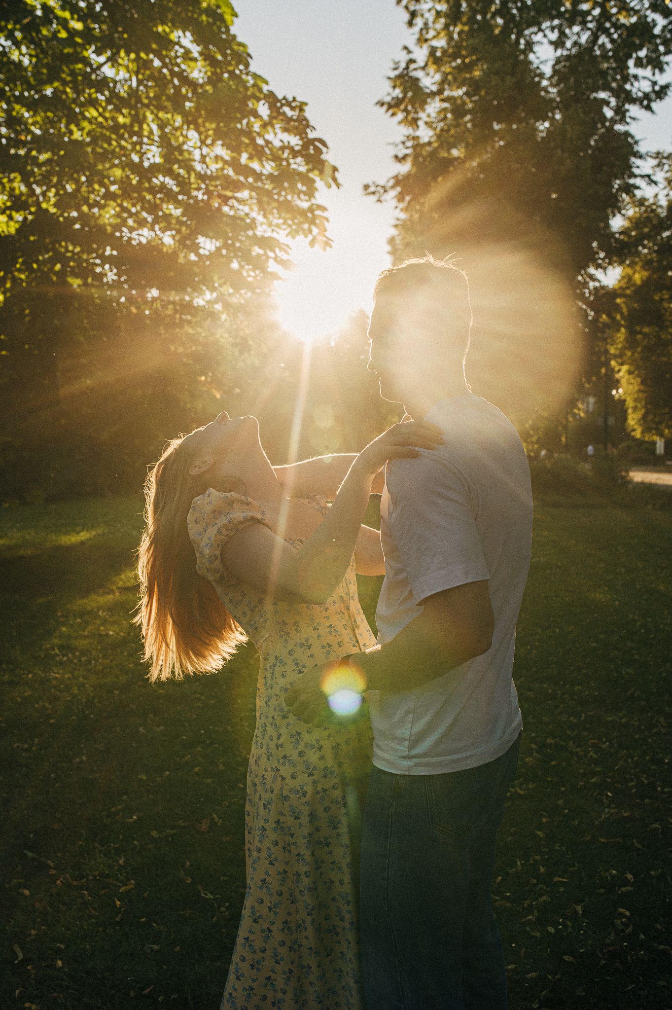 A romantic couple dancing together in a park during golden hour with sun flares.