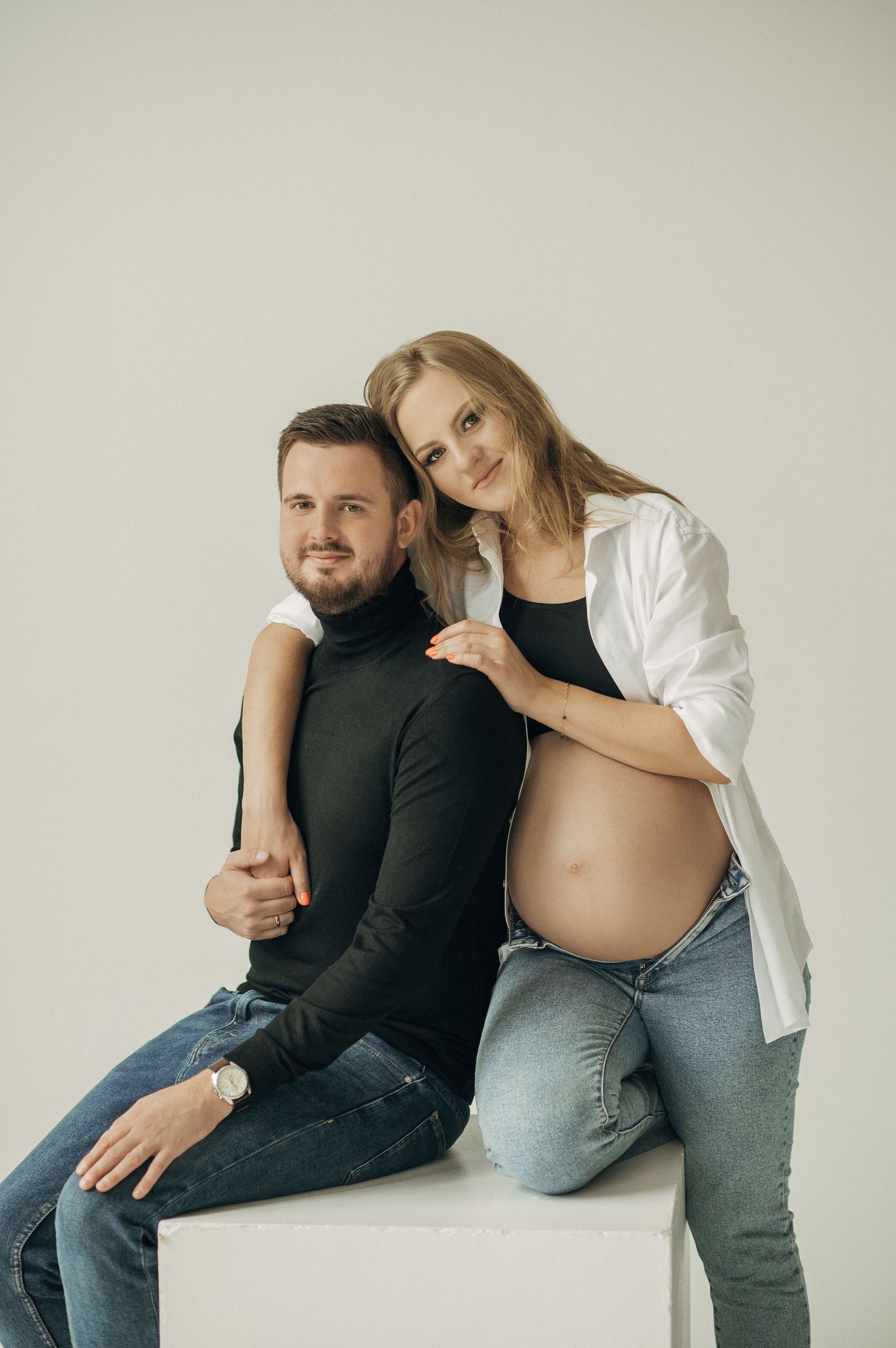 Intimate maternity photoshoot showing couple in casual clothing posing together against light background.