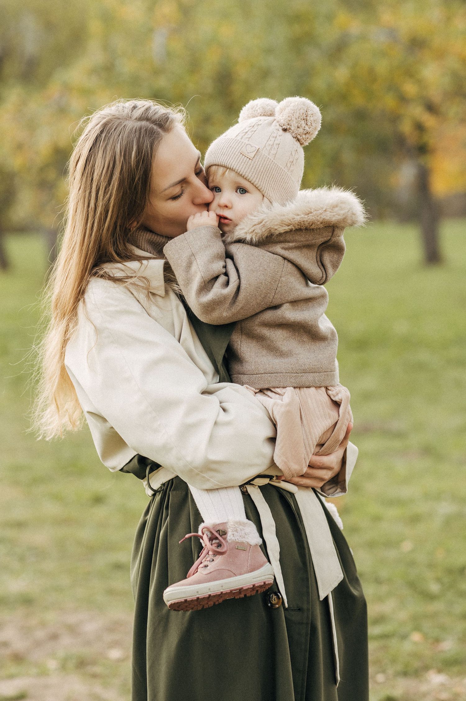 Parent and child share tender moments outdoors wearing matching beige outfits against a green natural backdrop.