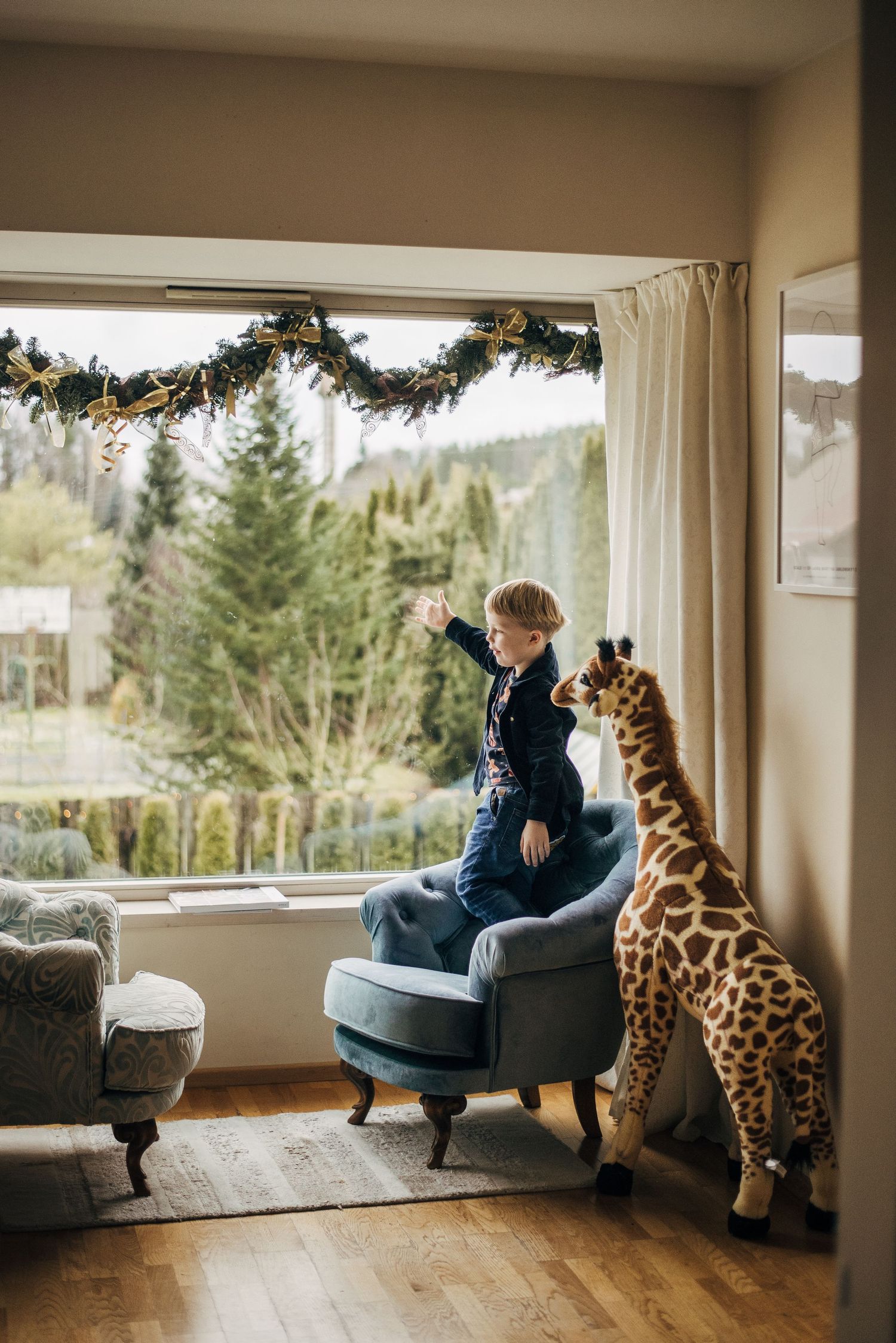 Child sitting in gray armchair by window with large stuffed giraffe toy.