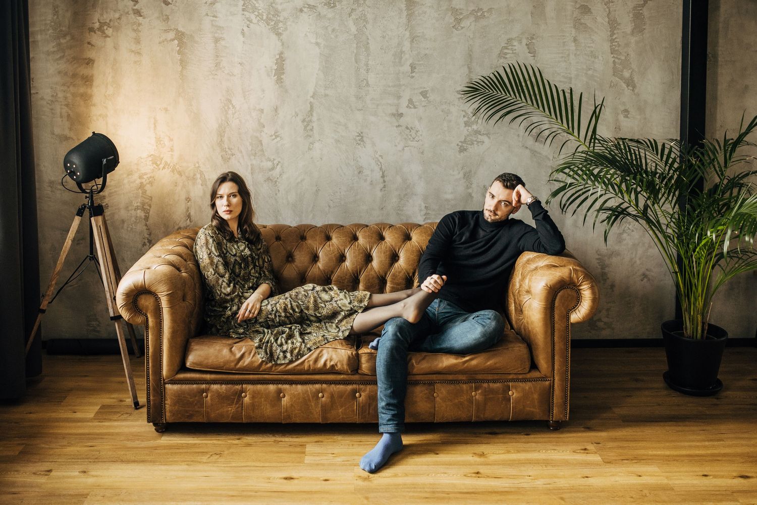 Two people relaxing on vintage brown leather Chesterfield sofa in modern industrial space.