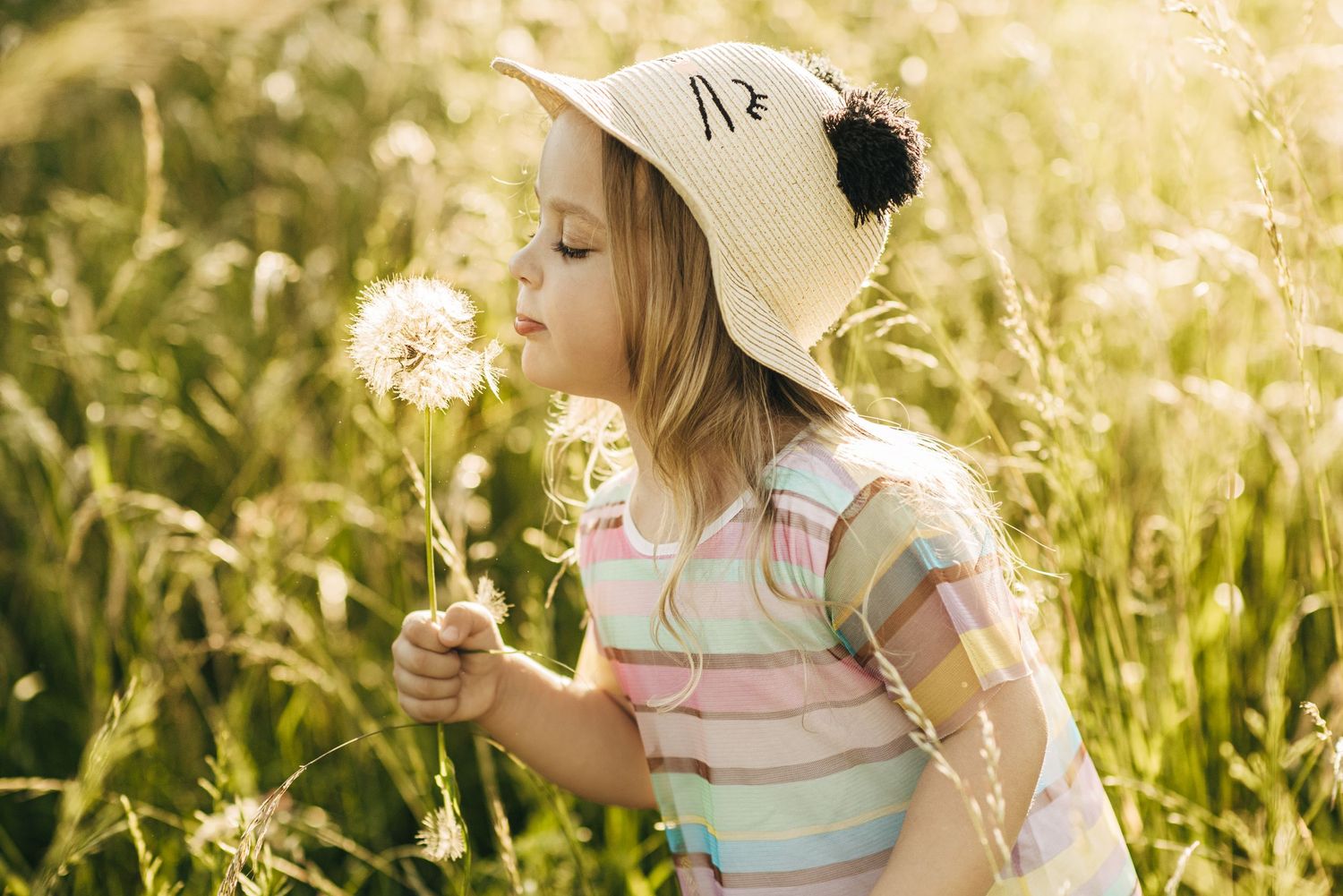 A child wearing a sun hat blows on a dandelion in a summer meadow.