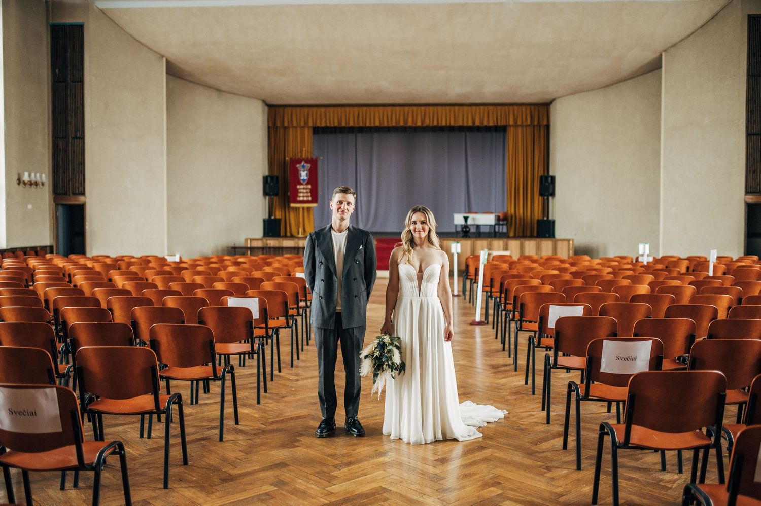 Wedding couple stands in empty auditorium with wooden chairs and stage.