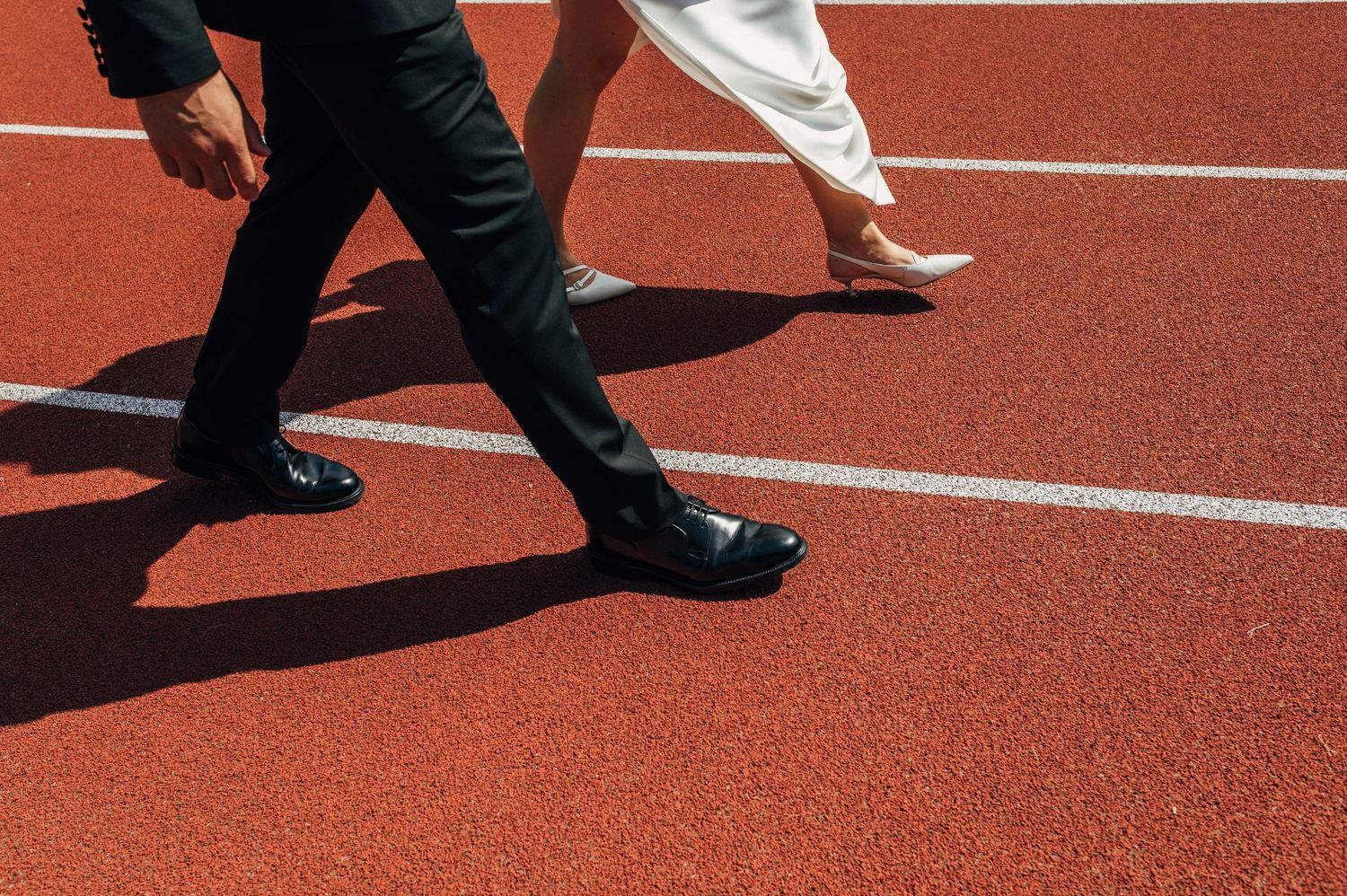 Two people in formal attire walking on a red running track, one in a suit and dress shoes, the other in a white dress.