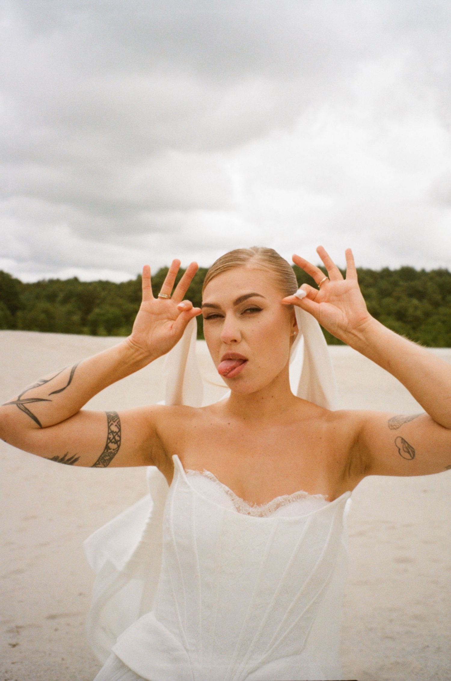 Tattooed bride in white strapless gown playfully sticking tongue out on sandy beach with cloudy sky.