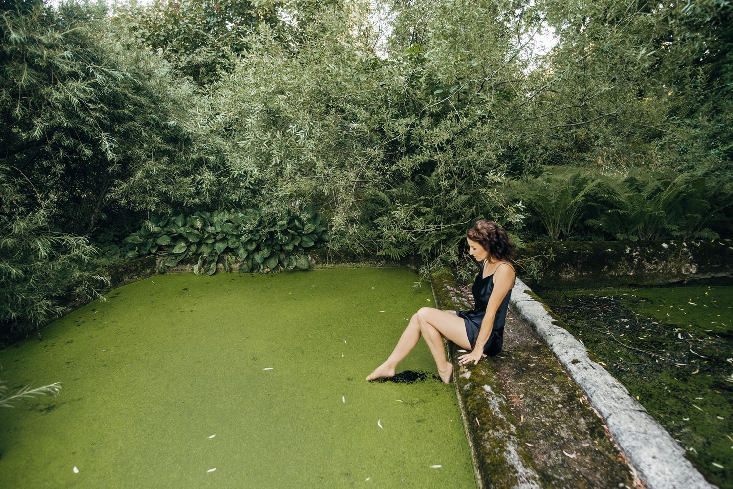 Figure relaxing on mossy stone wall near pond in lush green garden setting.