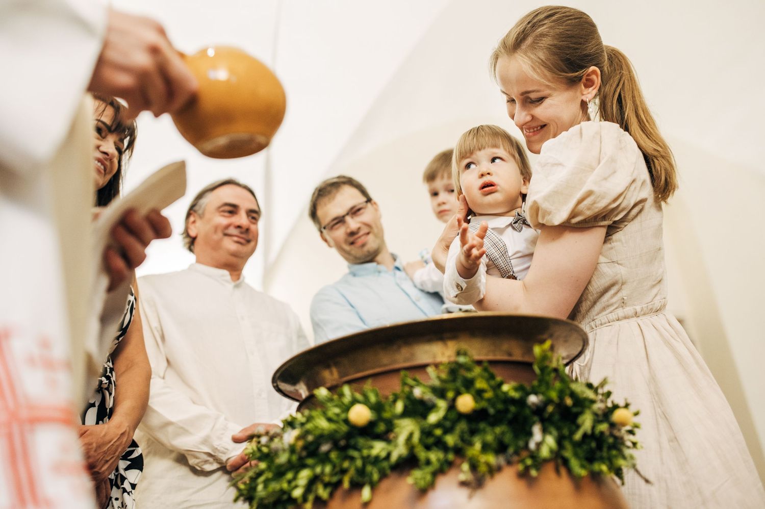 Priest performs baptism ceremony with family gathered around decorated font in church.