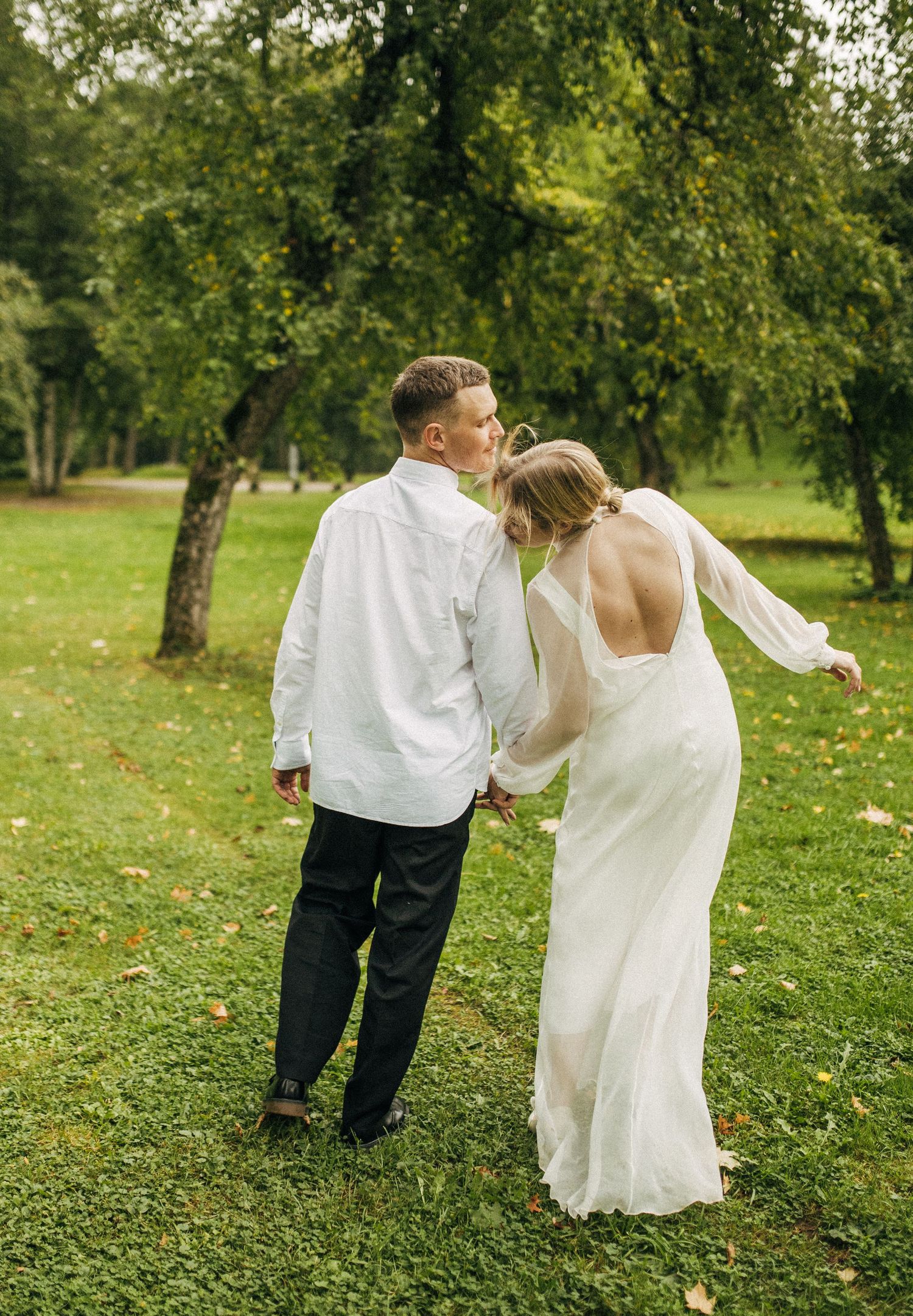 A couple in wedding attire walks together on a lush green lawn beneath trees during an outdoor celebration.