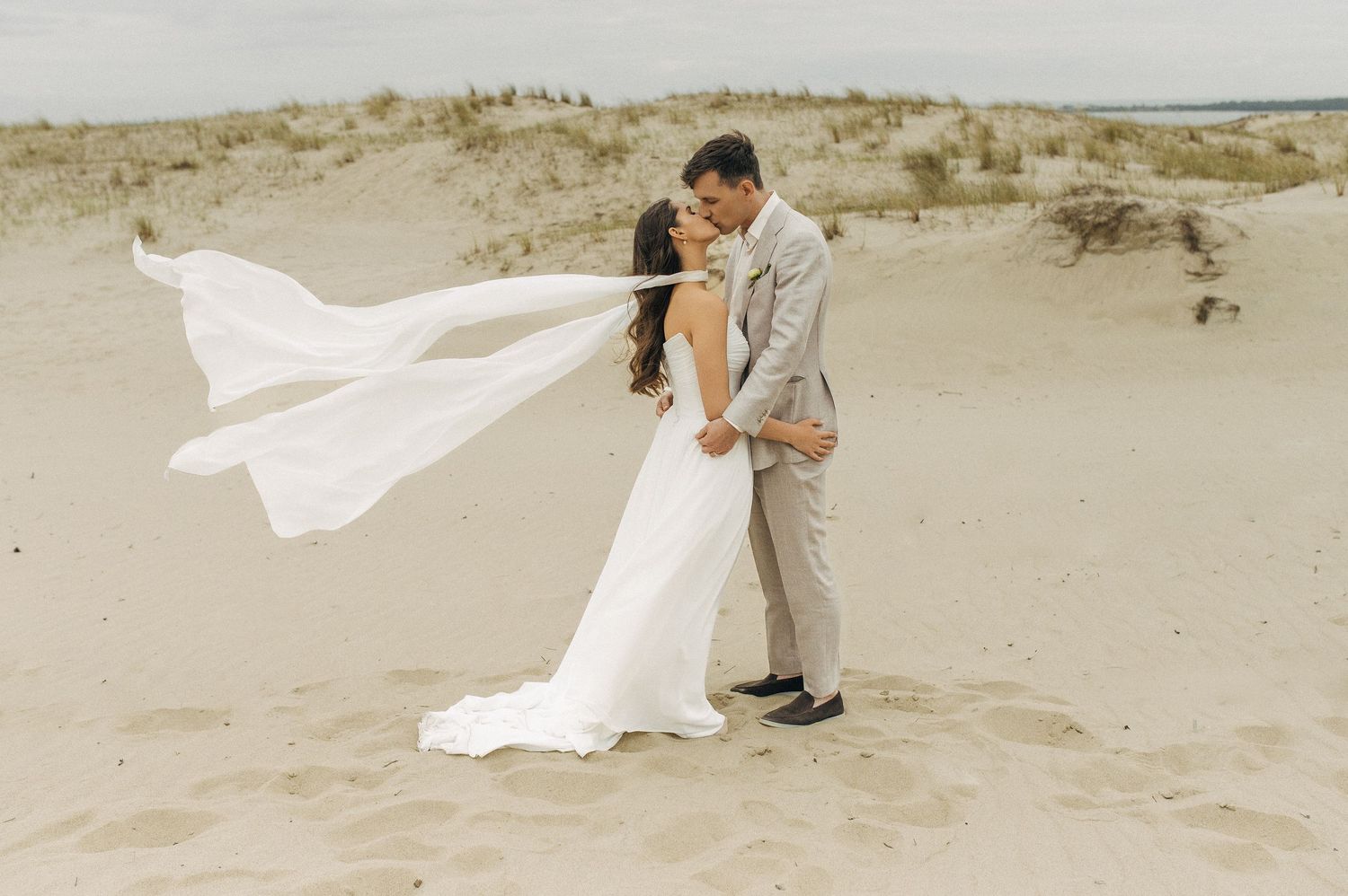 A couple embraces on a sandy beach while a white veil flows dramatically in the wind.
