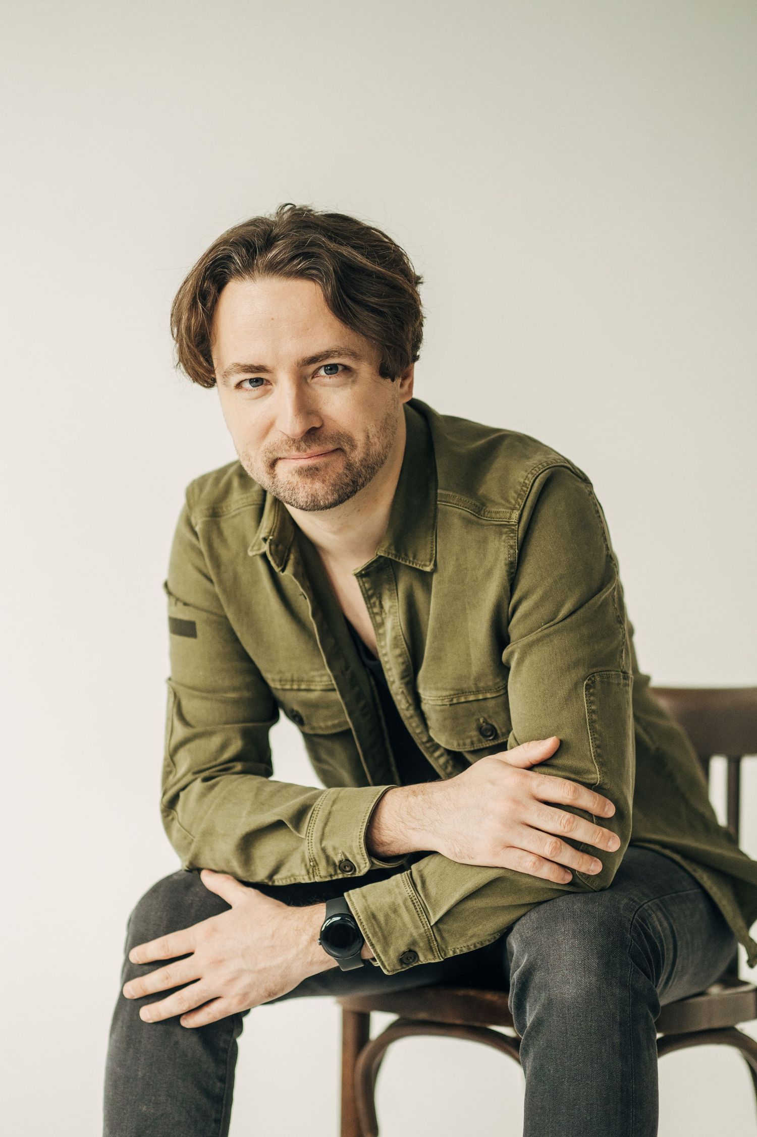 A person in an olive green jacket sits casually on a wooden chair against a white background in multiple poses.