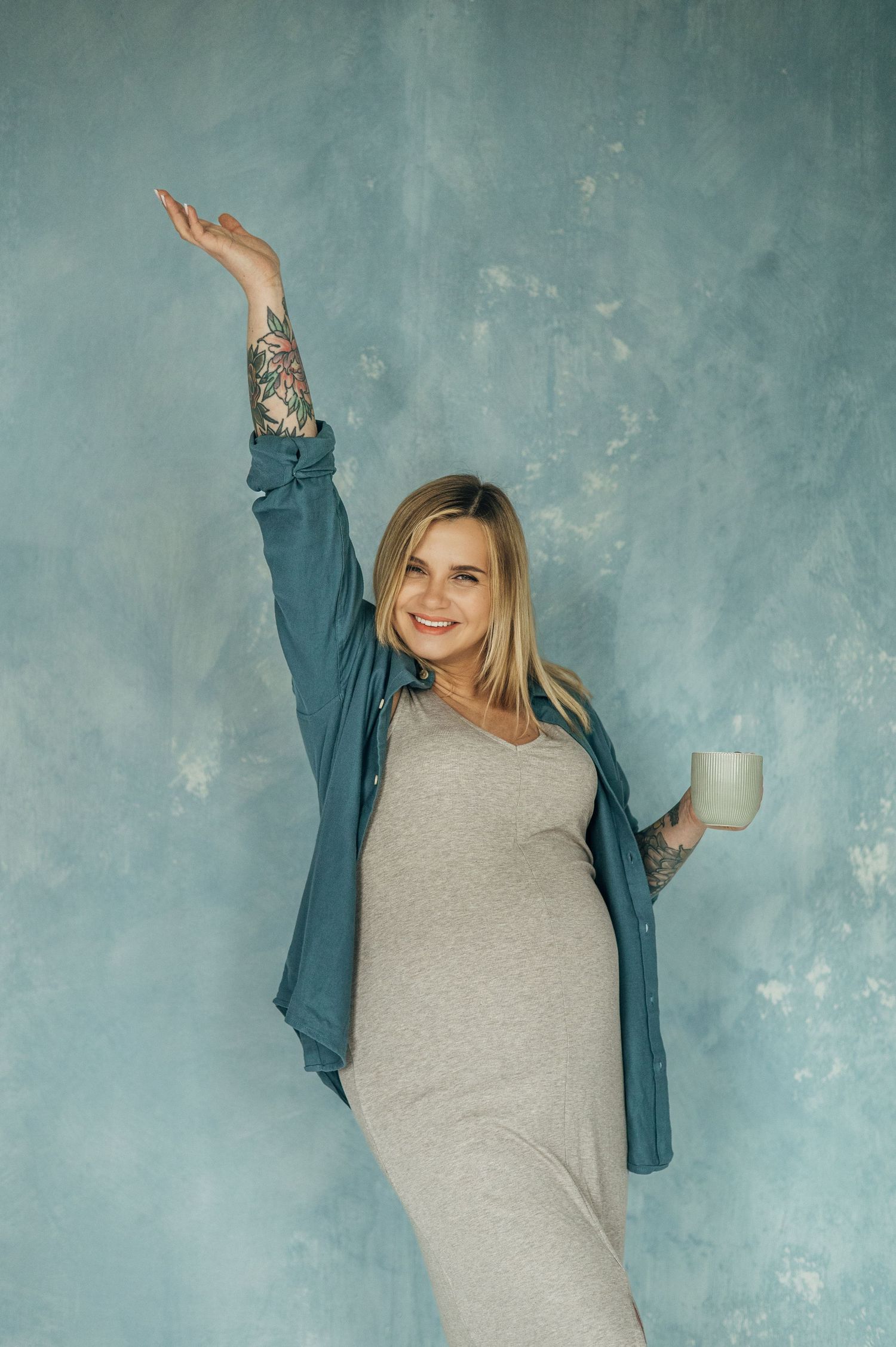 Pregnant woman in gray dress and blue cardigan poses joyfully with coffee mug against turquoise wall.
