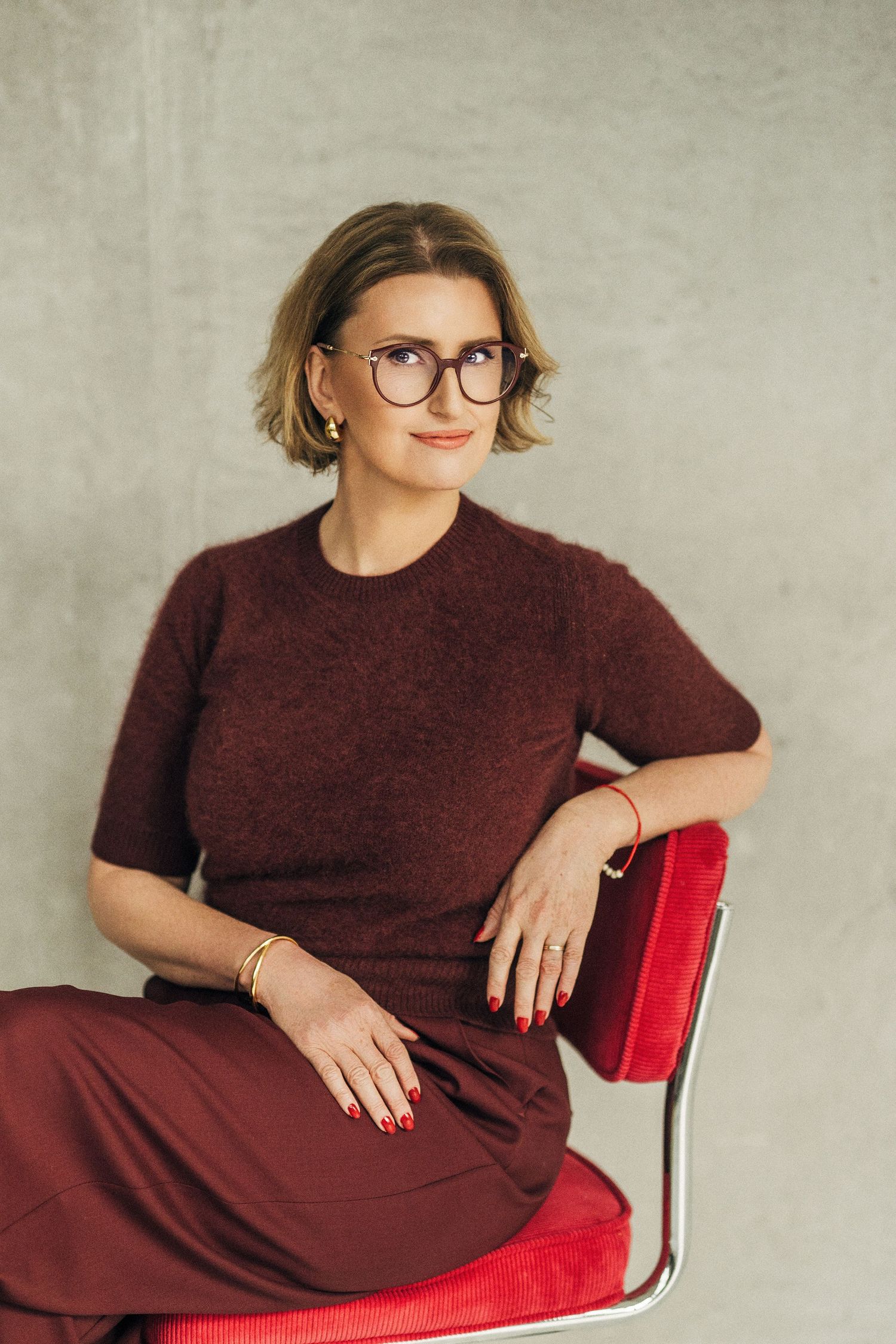 A figure in a burgundy dress sits on a bright red chair in a studio setting against a textured grey background.