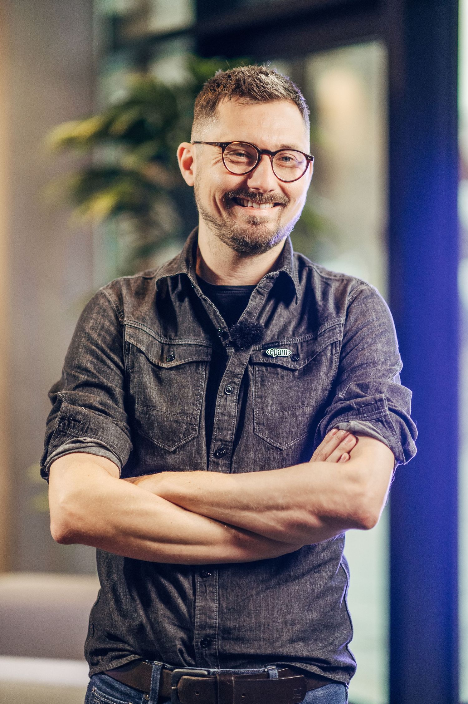 Professional wearing a dark denim shirt and glasses smiles warmly with arms crossed in an office setting.