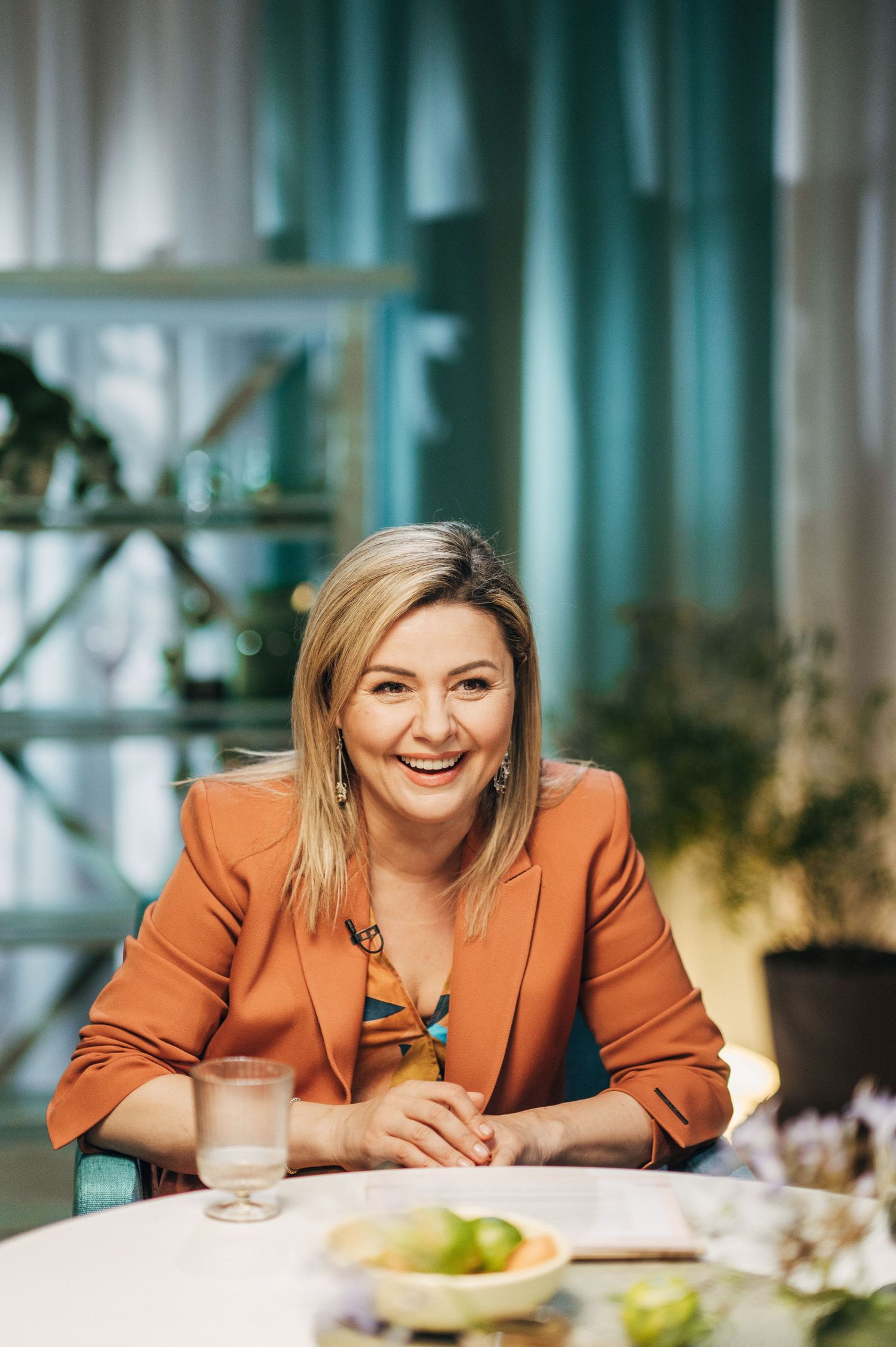 Professional wearing an orange blazer smiles warmly while seated at a modern dining table in an upscale setting.