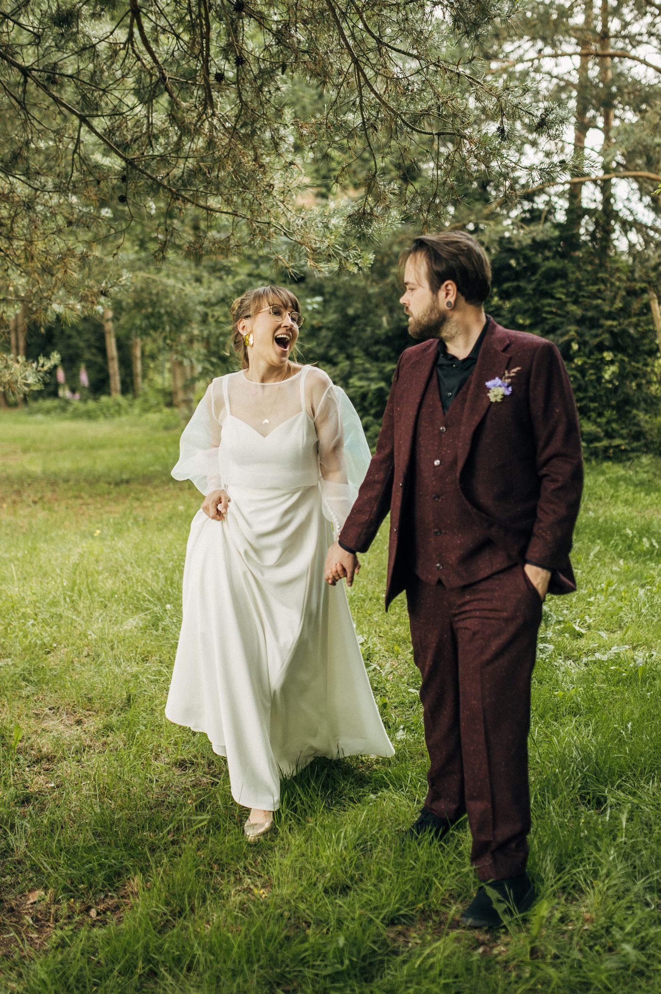 Newlyweds laughing together in sunlit meadow with burgundy and white attire.