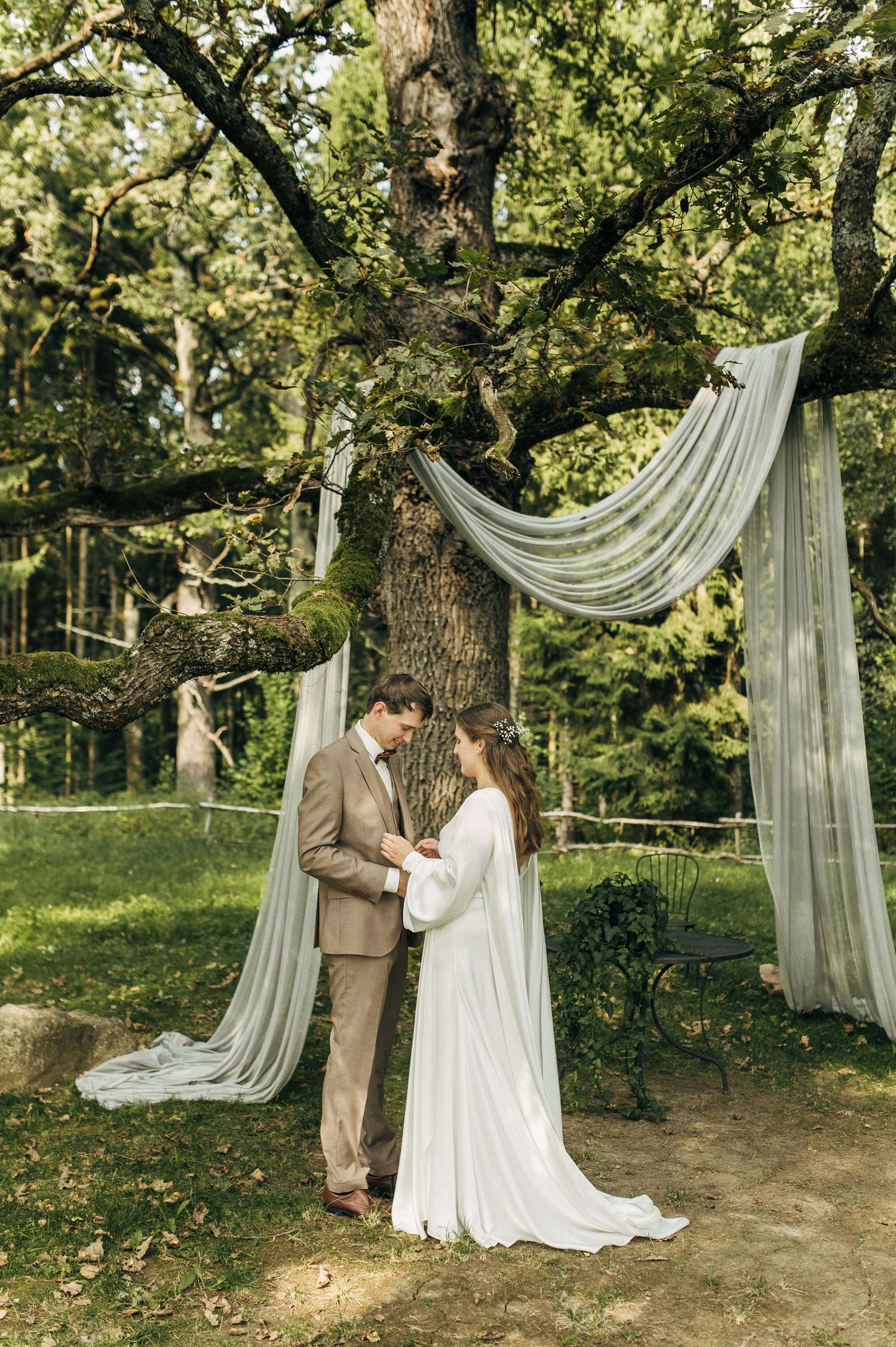 Elegant outdoor wedding ceremony setup with flowing white drapes between trees and a couple sharing a romantic moment.