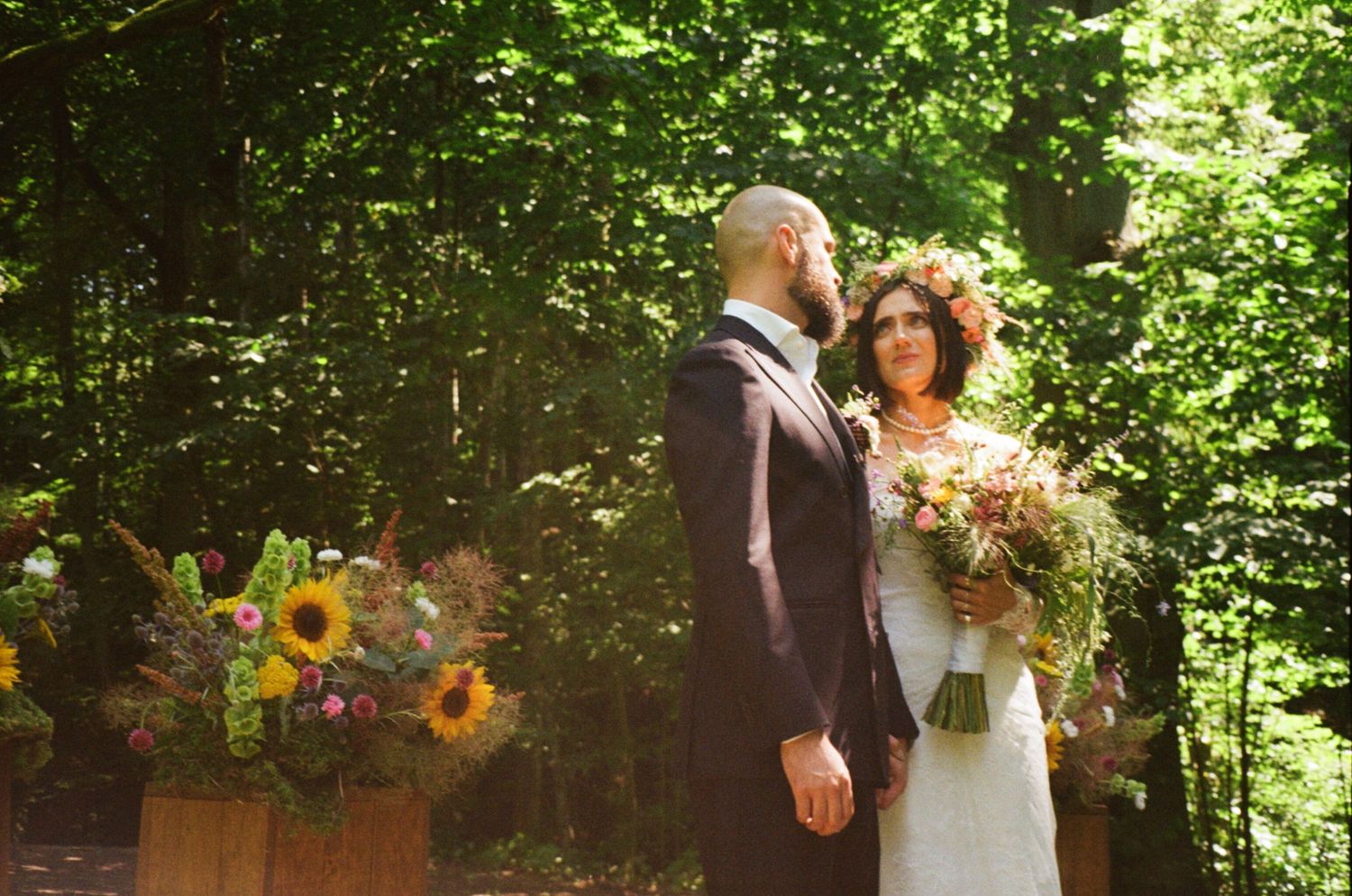 Bride holding sunflower bouquet stands with groom in suit amid lush green forest backdrop at outdoor wedding ceremony.