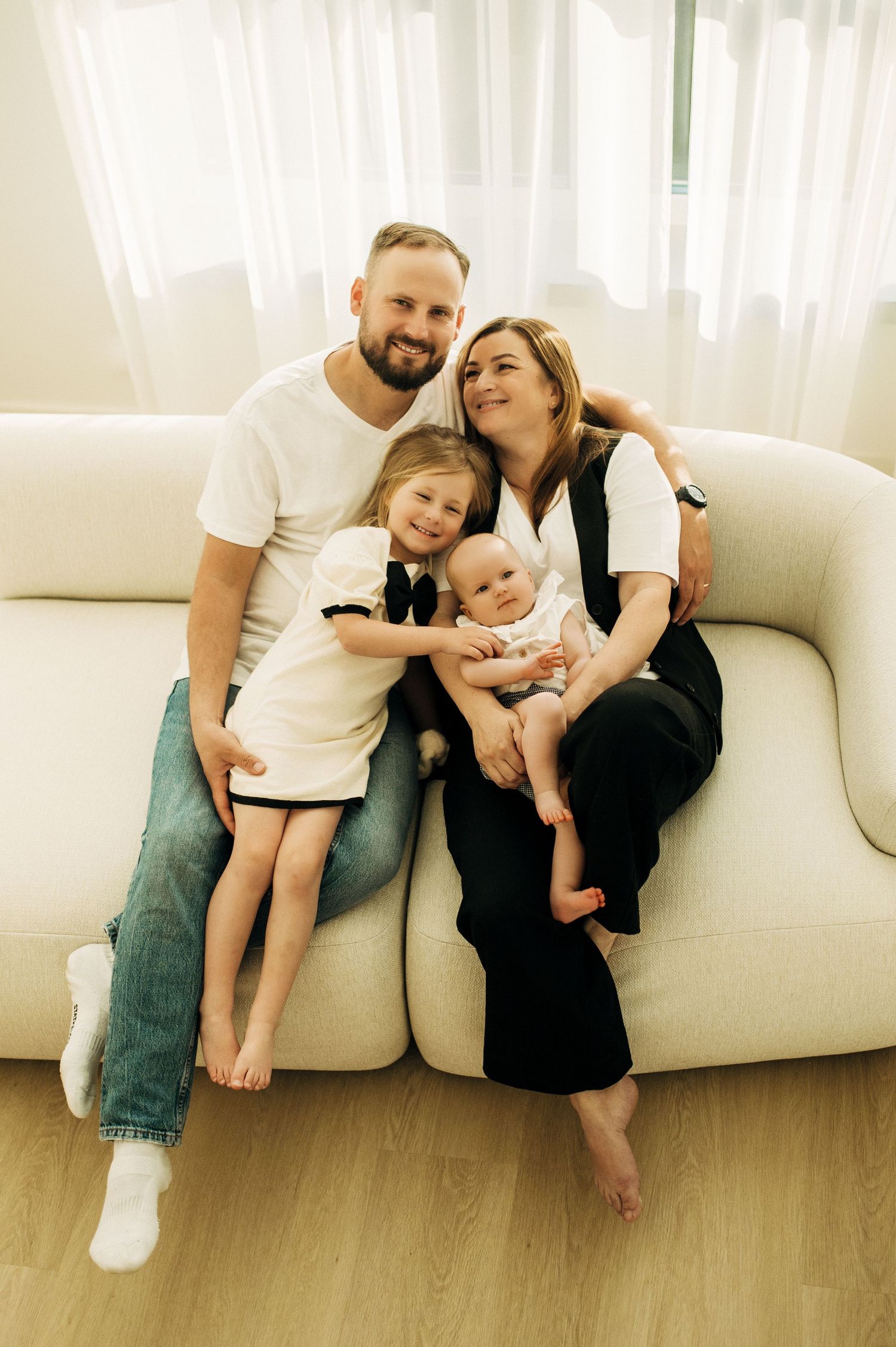 Family relaxing together on white couch in bright room with wooden floors.