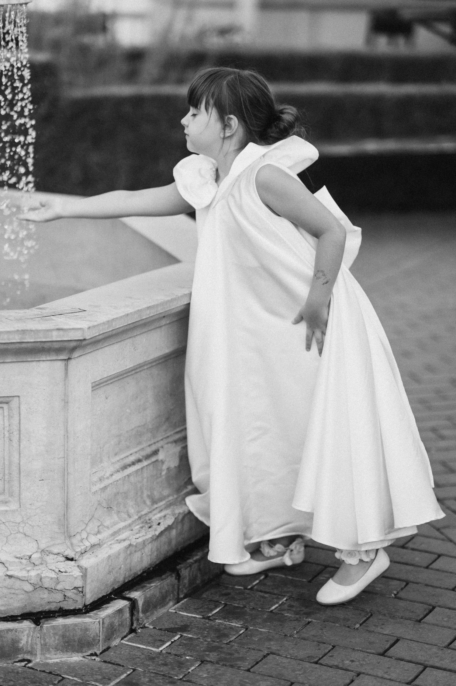 A young girl in a white dress reaches into a stone fountain, captured in elegant black and white photography.