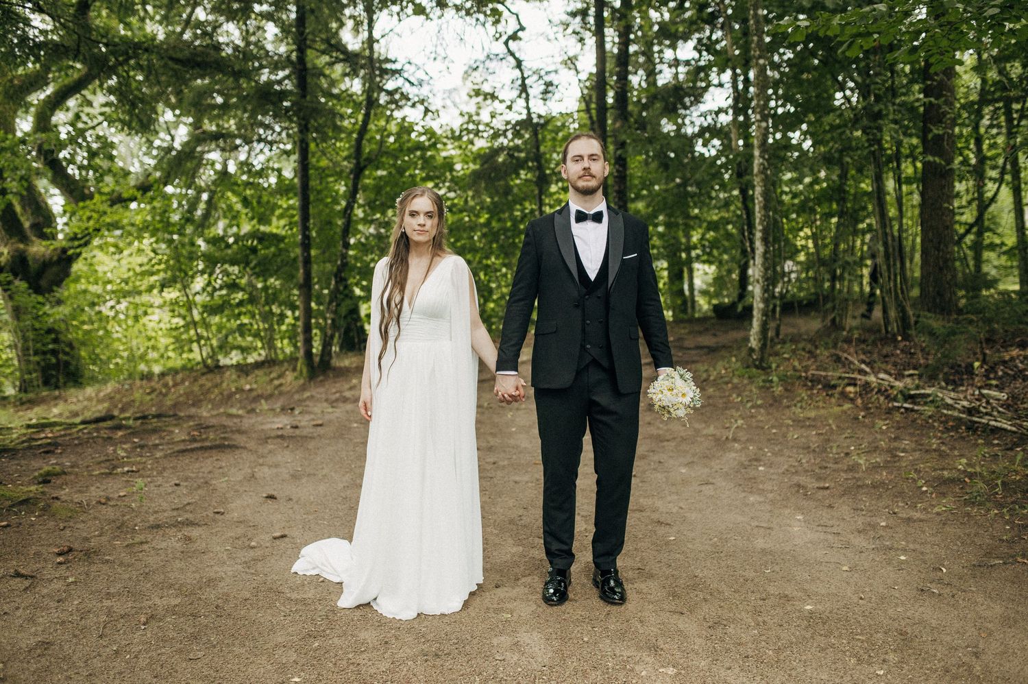 A couple in formal wedding attire poses on a forest path surrounded by greenery.