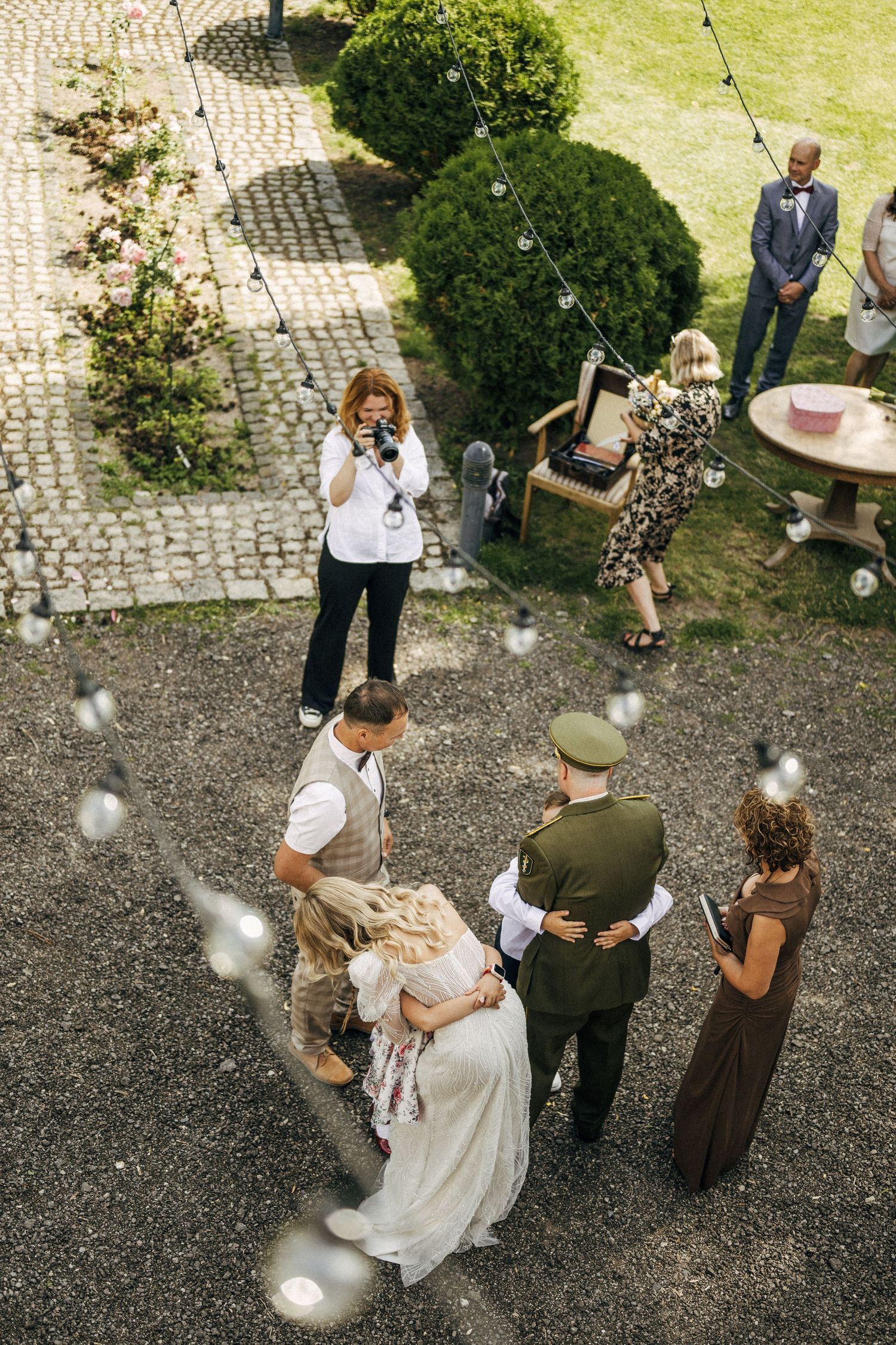 Aerial view of wedding party walking on cobblestone path with hanging string lights.