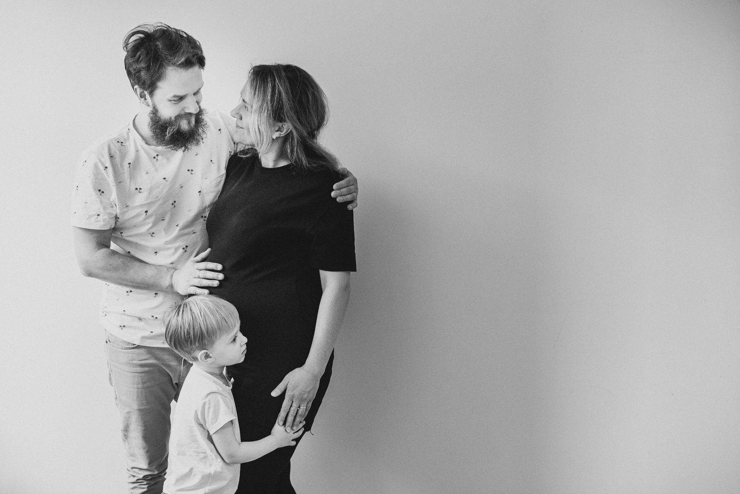 Black and white portrait of a family of three sharing an intimate moment against a plain wall.