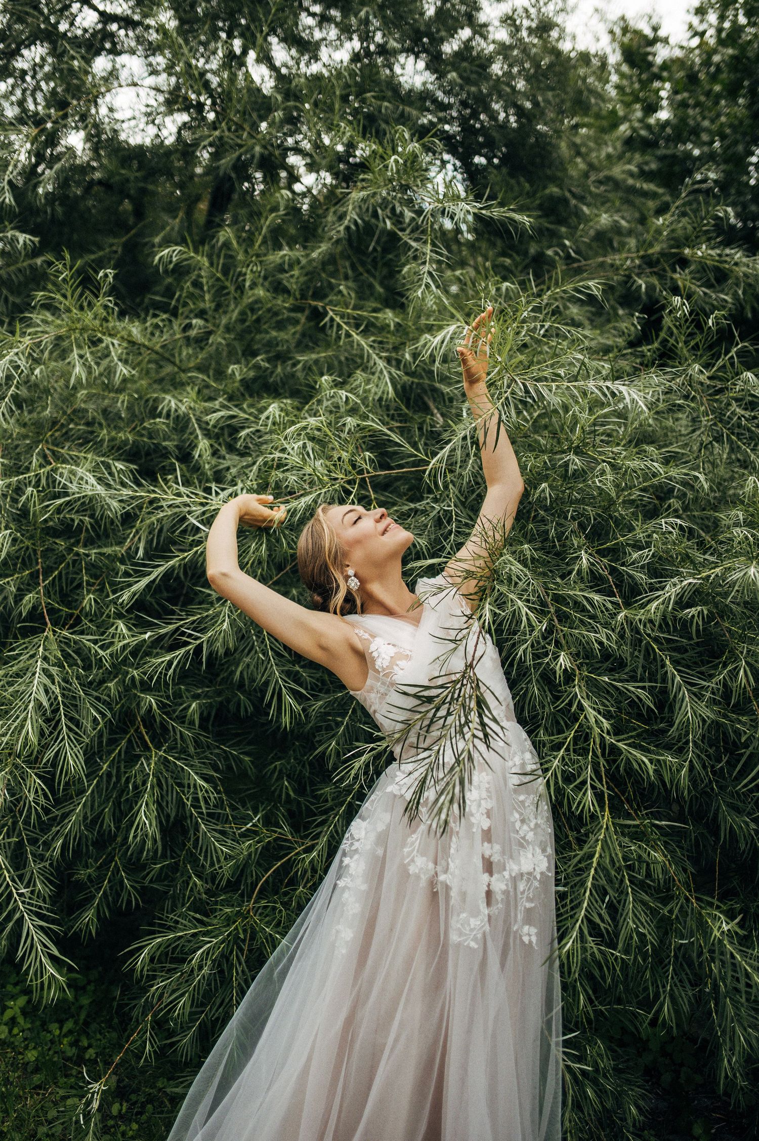 A person in a flowing white tulle gown reaches up gracefully among evergreen branches in a dreamy outdoor setting.