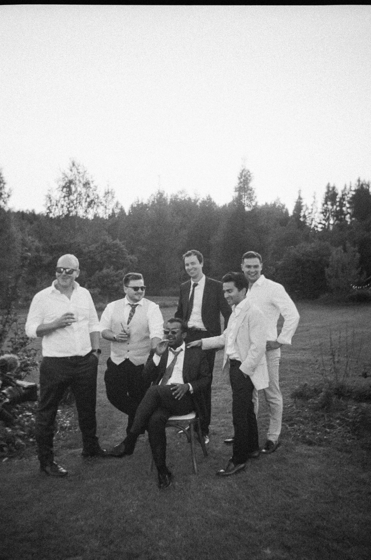 A group of men in formal attire pose playfully on a grassy hill in black and white.