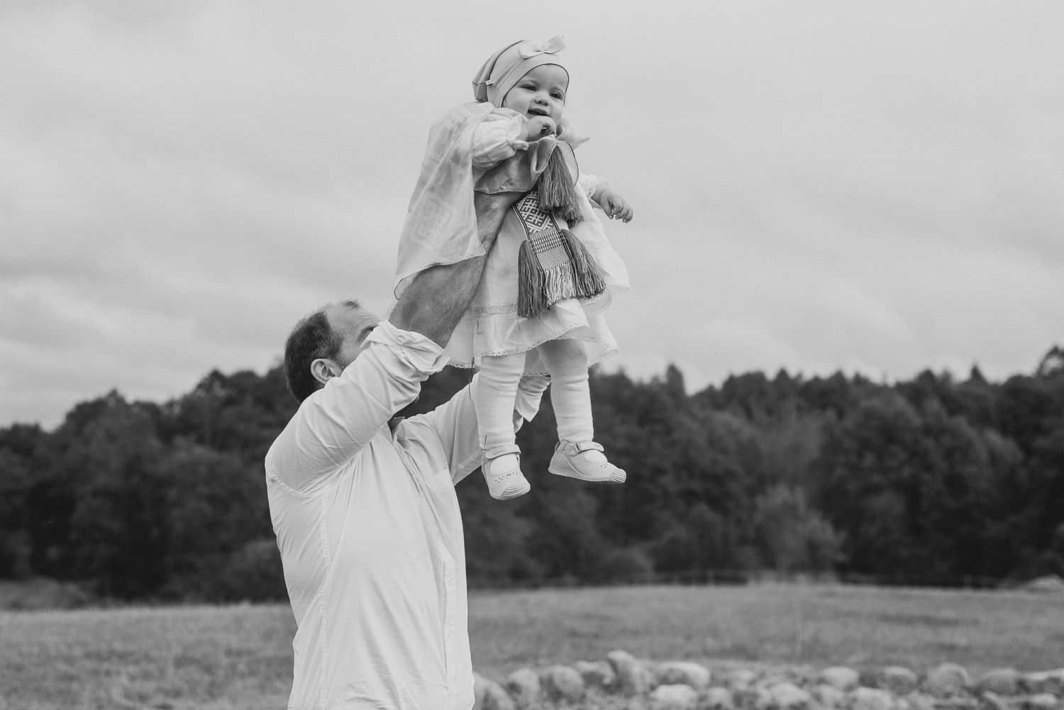 Black and white photo of father lifting baby girl above his head in an open field.